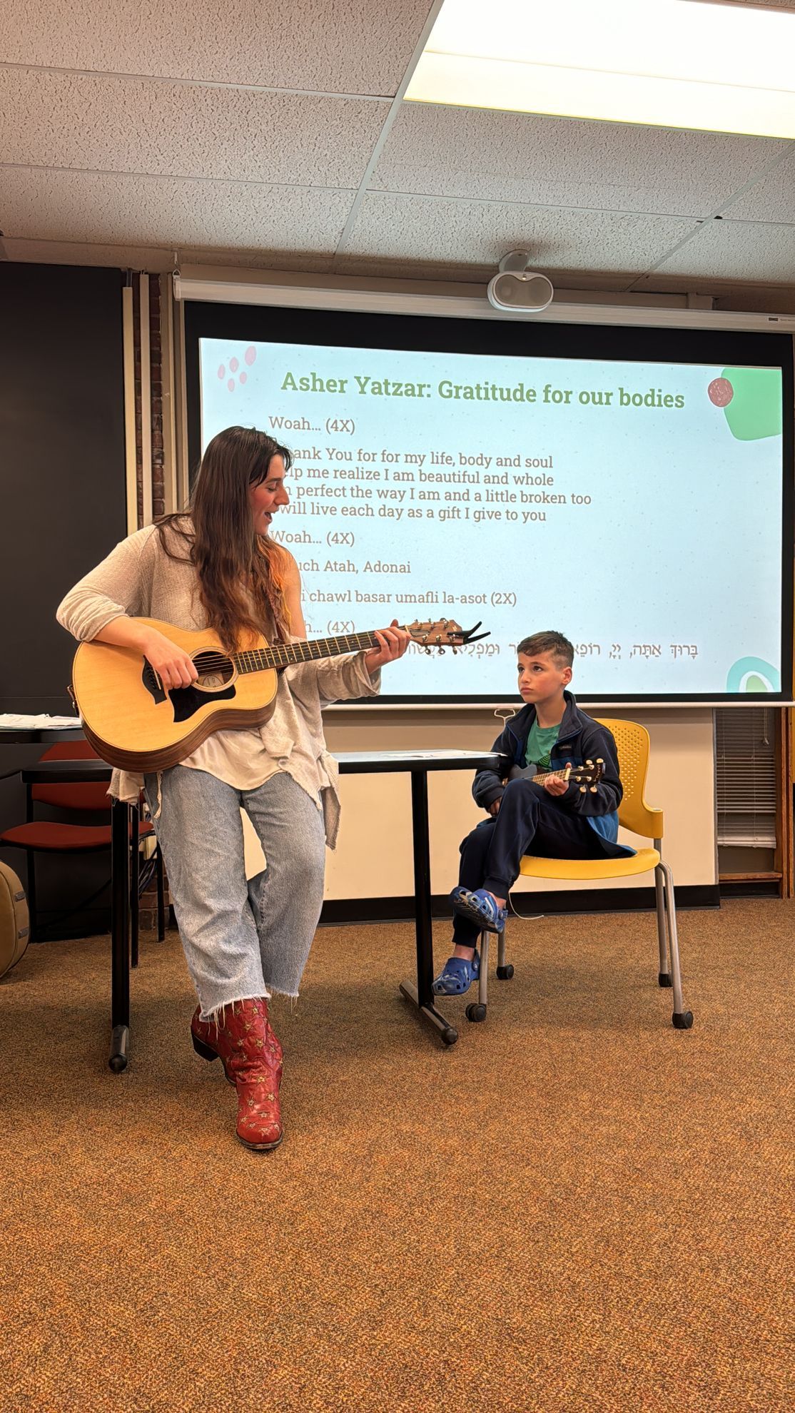 A woman is playing a guitar in front of a boy in a classroom.