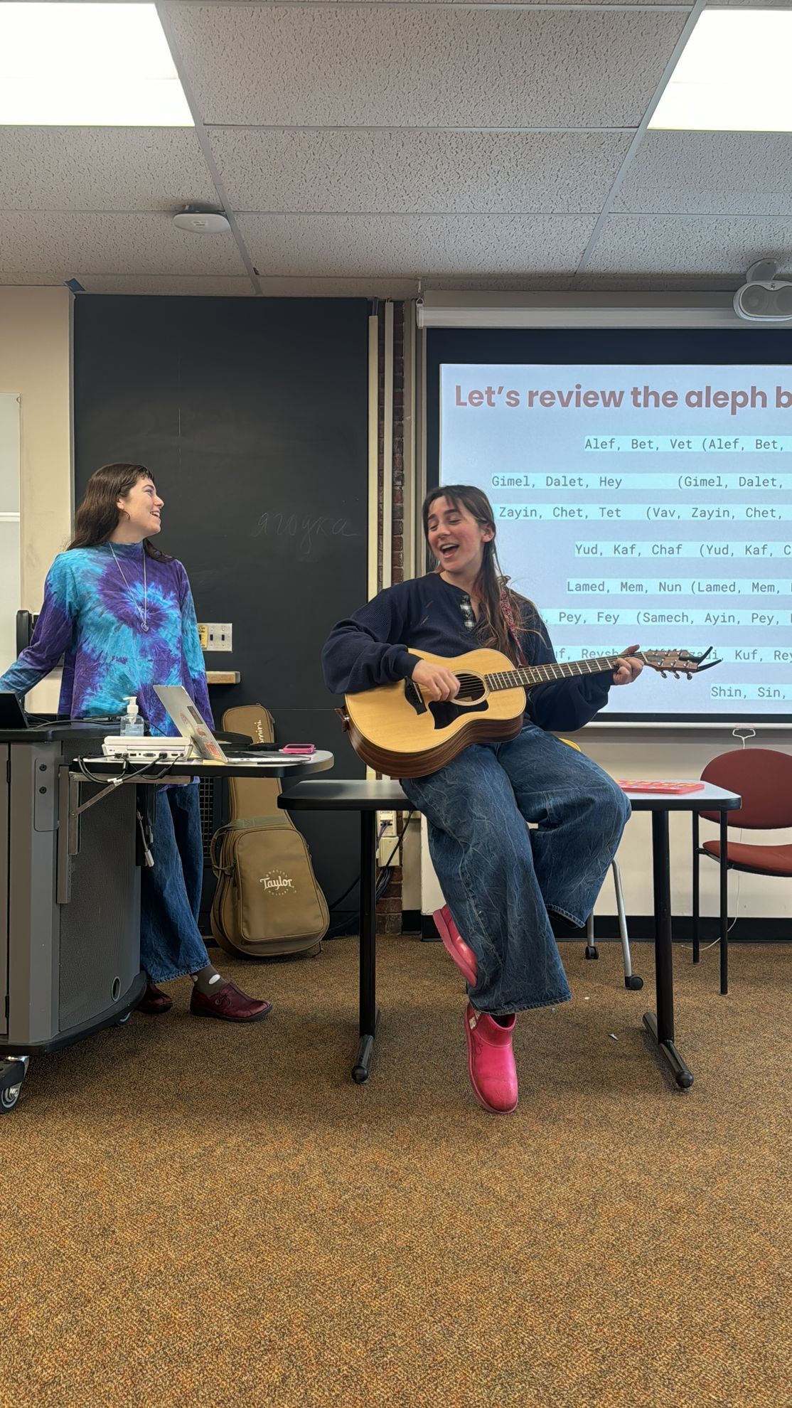 A woman is sitting at a table playing a guitar in a classroom.