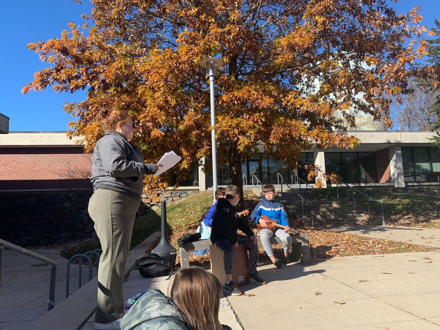 A woman is standing in front of a group of people sitting under a tree.