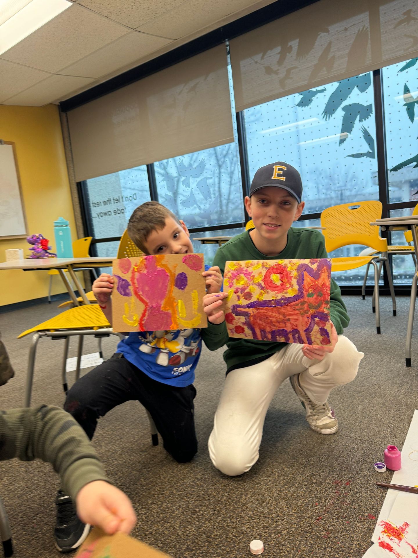 A boy with the letter e on his hat is kneeling next to a boy holding a painting