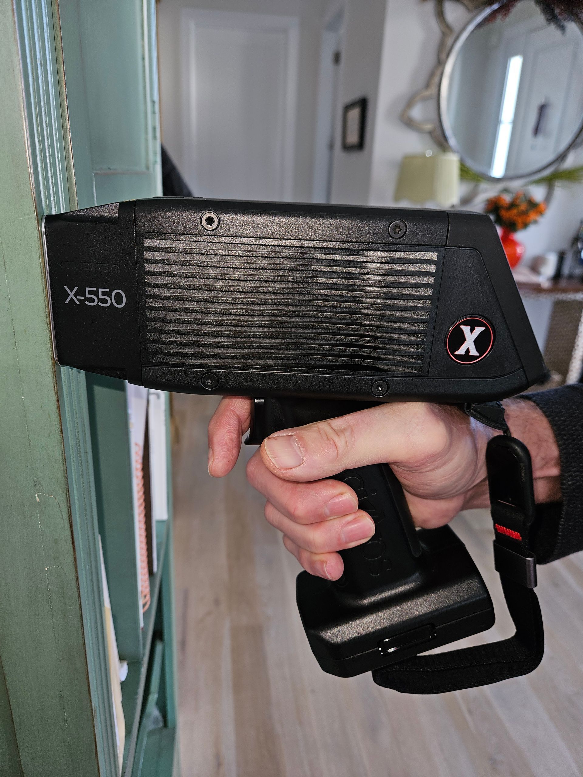 Hand holding an X-550 handheld device, pointed at a bookcase. The device is black with an 