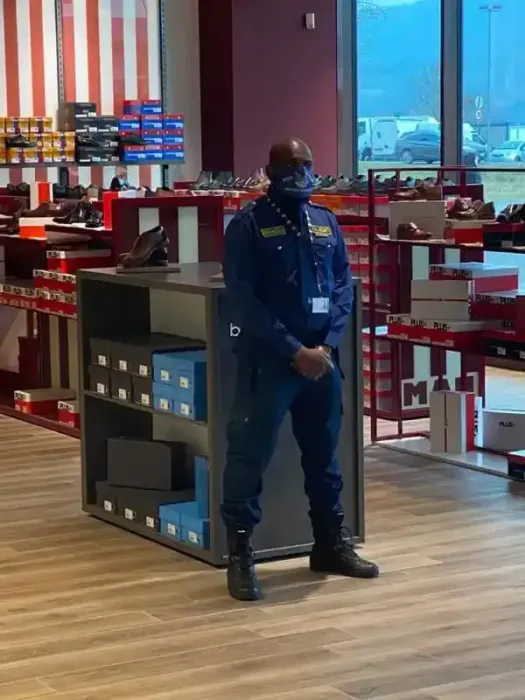 Una guardia di sicurezza si trova all'interno di un negozio, vicino a scaffali espositivi rossi, e indossa un'uniforme scura e una mascherina.