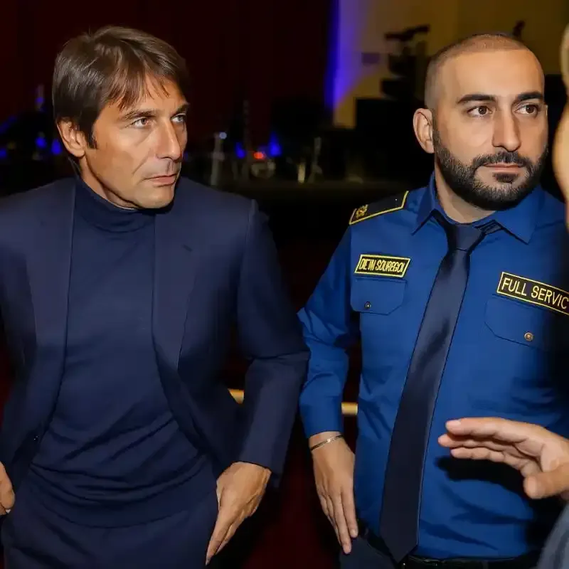 Due uomini in abiti blu scuro camminano all'interno di un edificio, uno con una giacca blu scuro e l'altro con un'uniforme blu da guardia di sicurezza.