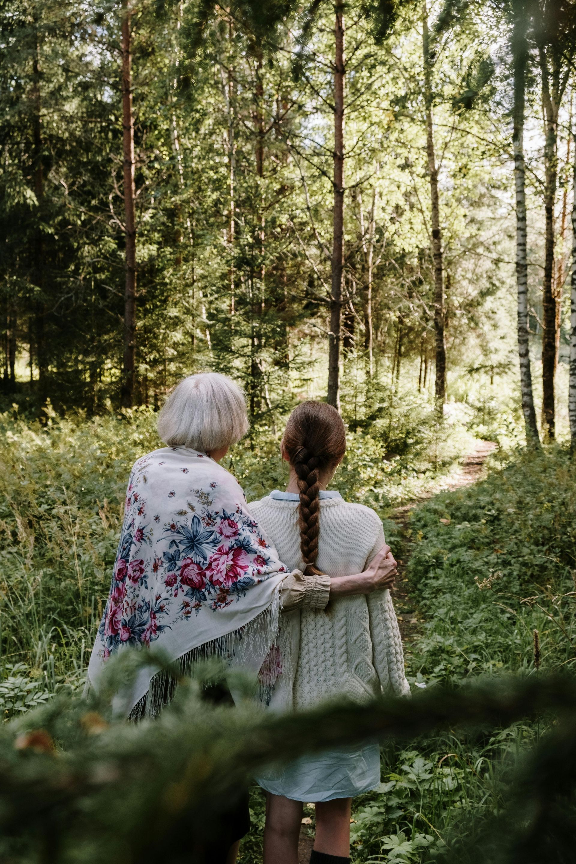 Two people standing with arms around each other in a sunlit forest, viewed from behind.