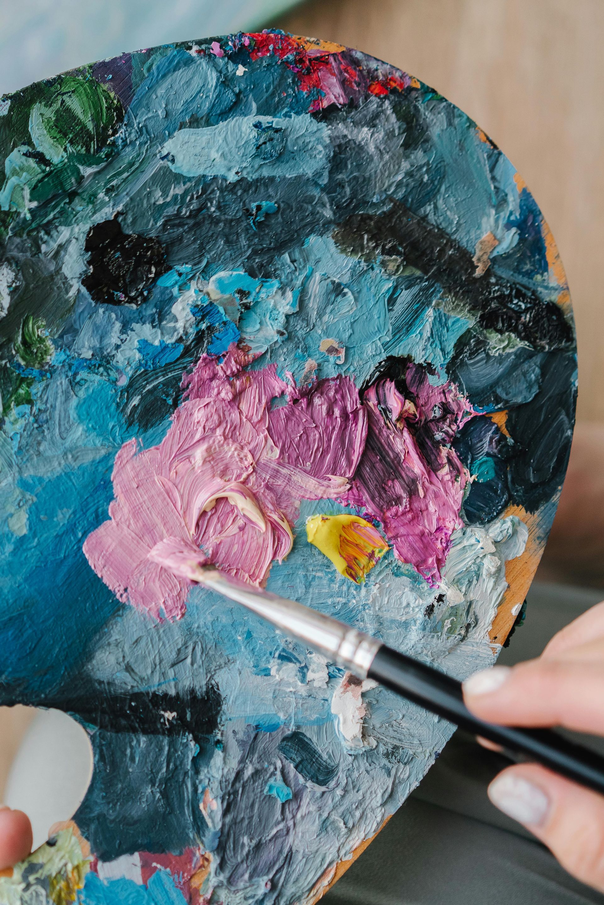 An artist's hand mixes pink oil paint on a textured palette using a small, fine-tipped paintbrush.