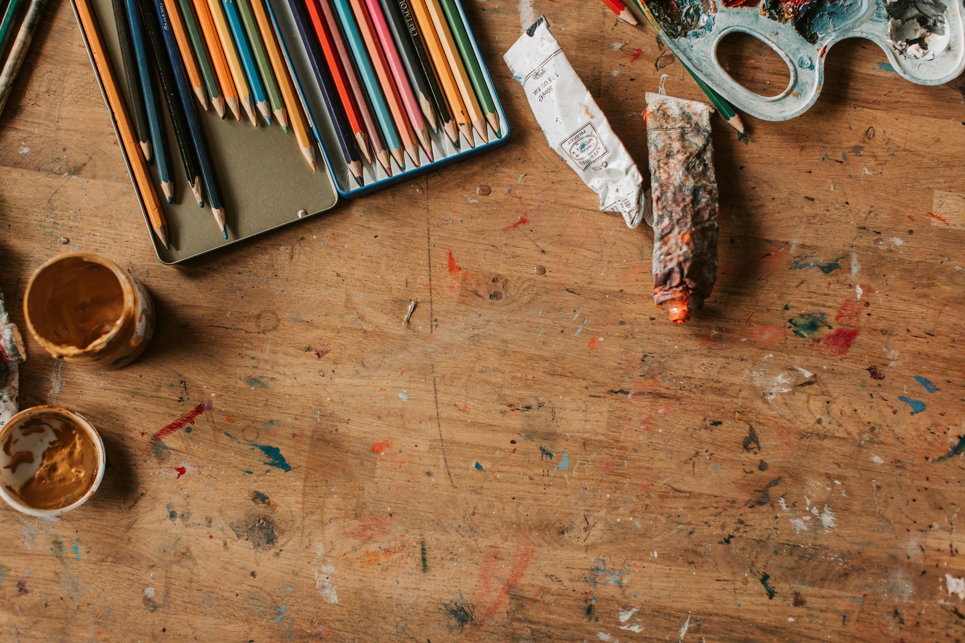 A wooden art desk with a box of colorful pencils, two small paint pots, a paint tube, and a palette.