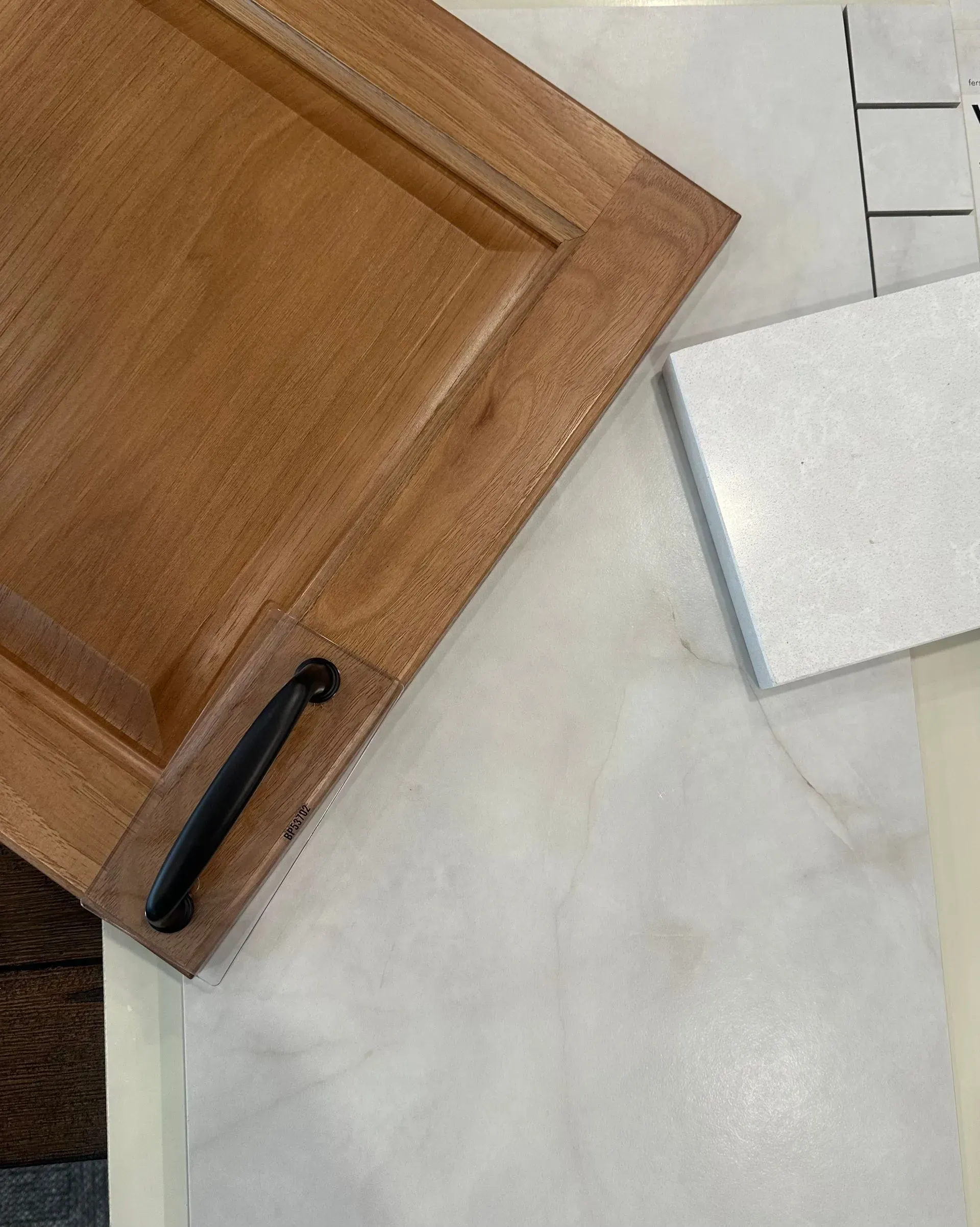 Wooden cabinet door with black handle next to a white countertop, white square, and tile samples.