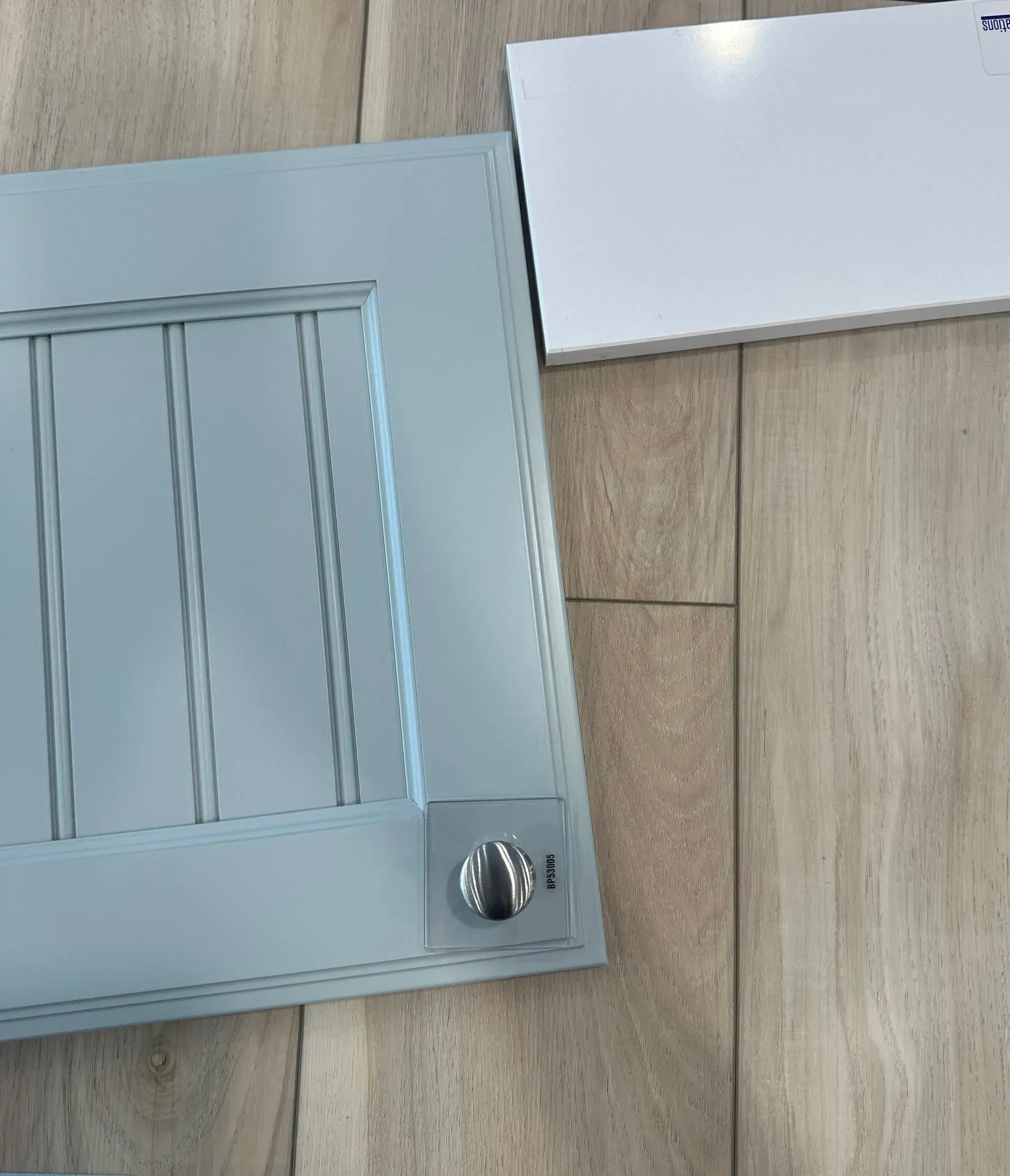 Light blue cabinet door with vertical slats and silver knob next to a white countertop sample on wood-look surface.