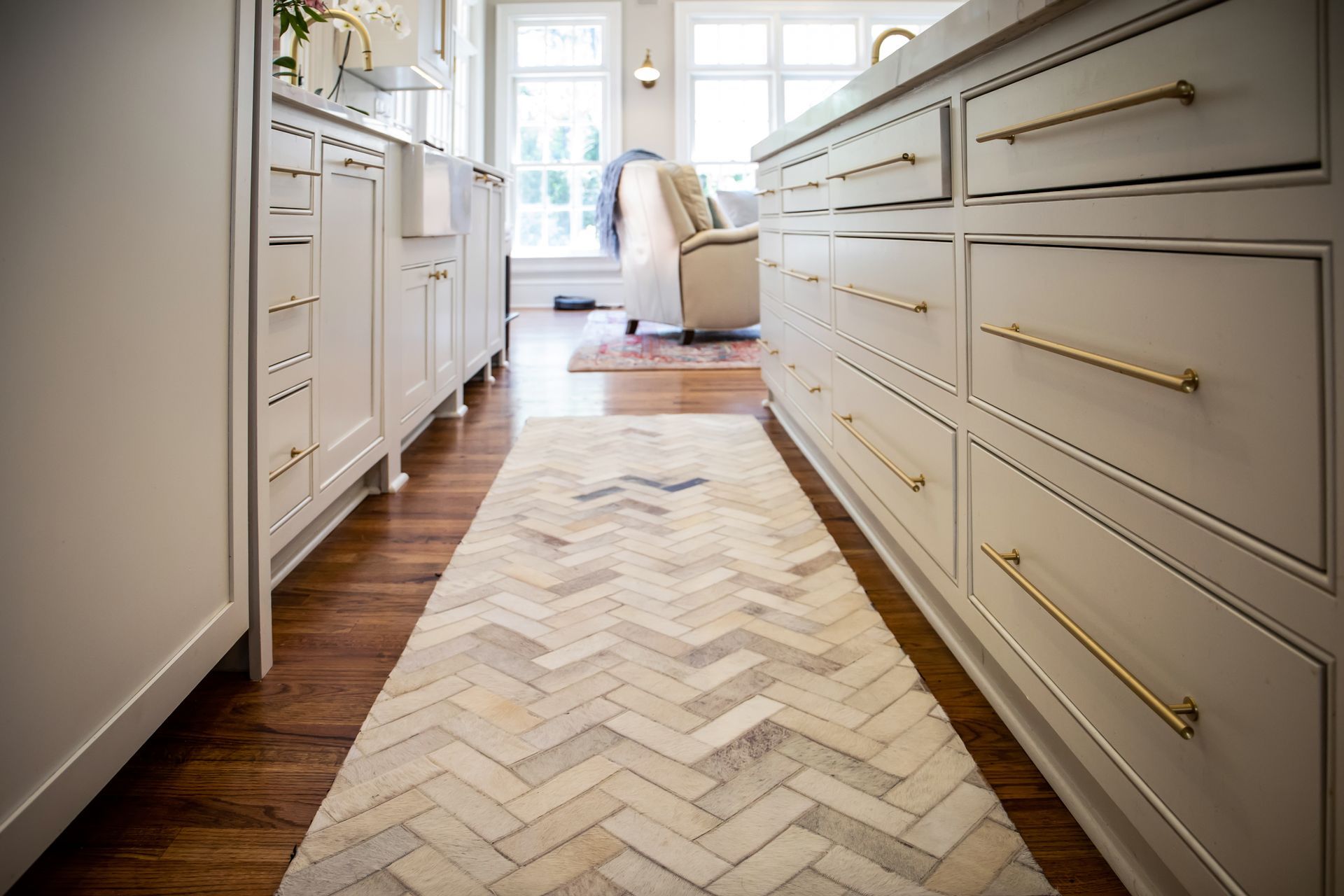 Kitchen with a light-colored herringbone rug, white cabinets with gold hardware, and a view to a sunny window.
