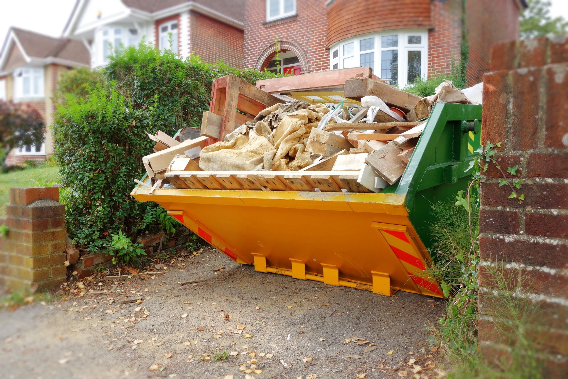 A yellow dumpster filled with junk is parked in front of a brick house.