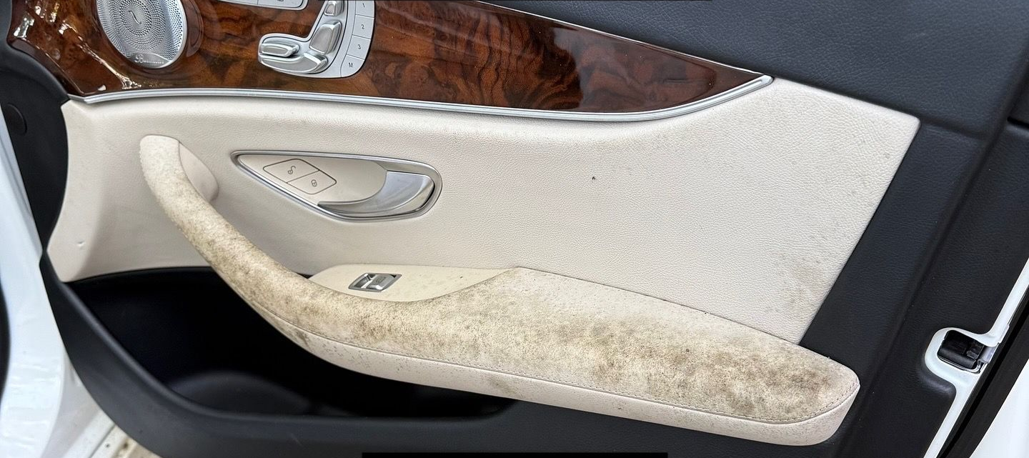 Close-up of a luxury car door panel featuring light beige upholstery, wood trim, and a heavily soiled, stained armrest.