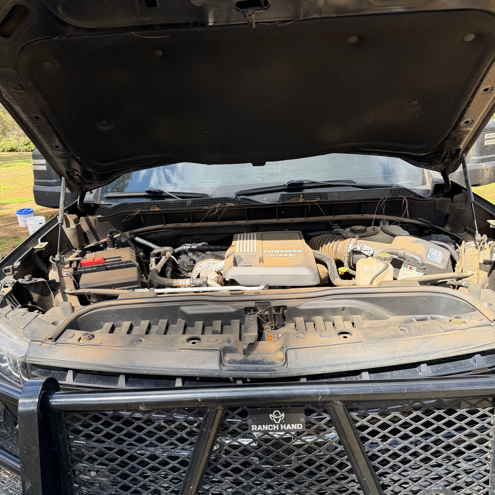 A close-up of an open truck hood revealing a dusty vehicle engine bay with a metal grille guard in the foreground.