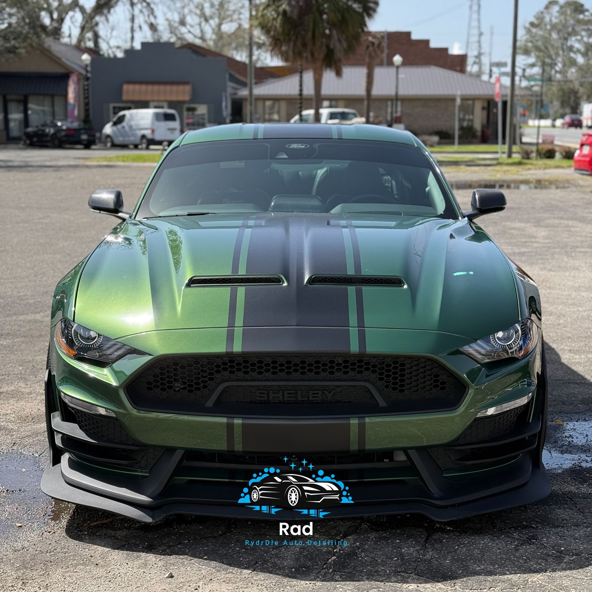 A green Ford Mustang with black racing stripes parked on a paved lot in front of buildings.