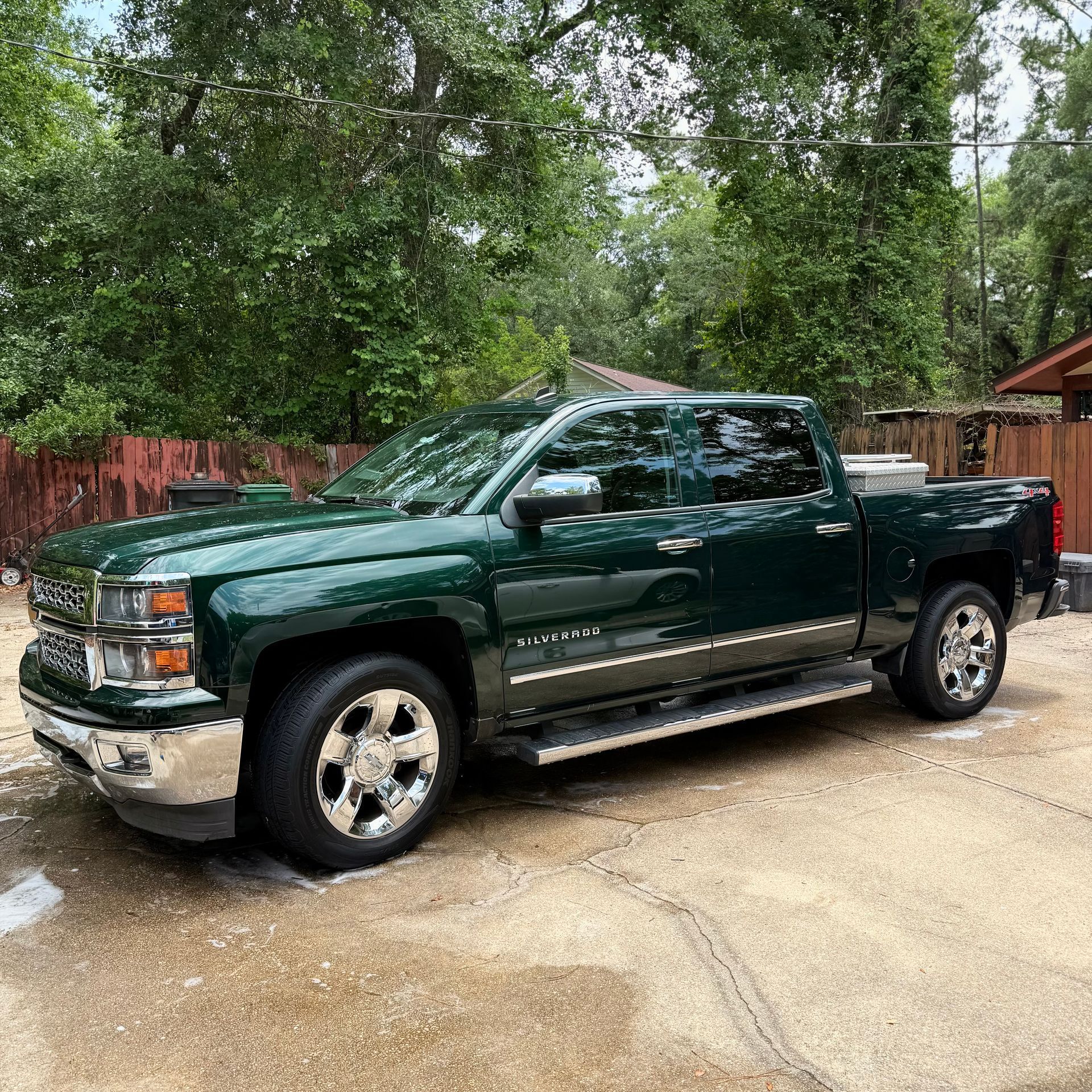 A dark green Chevrolet Silverado crew cab pickup truck parked on a concrete driveway with trees in the background.