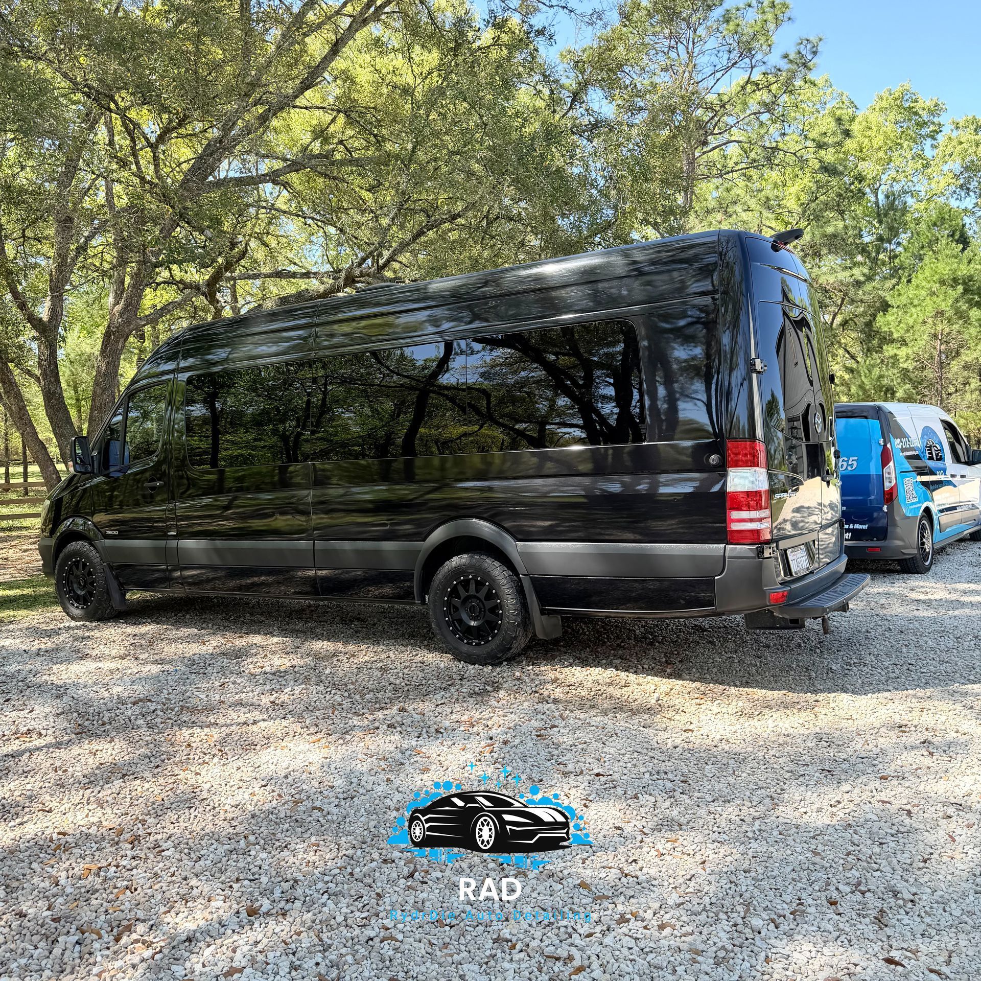 A black high-roof Sprinter van parked on a gravel lot next to a blue and white utility van under trees.