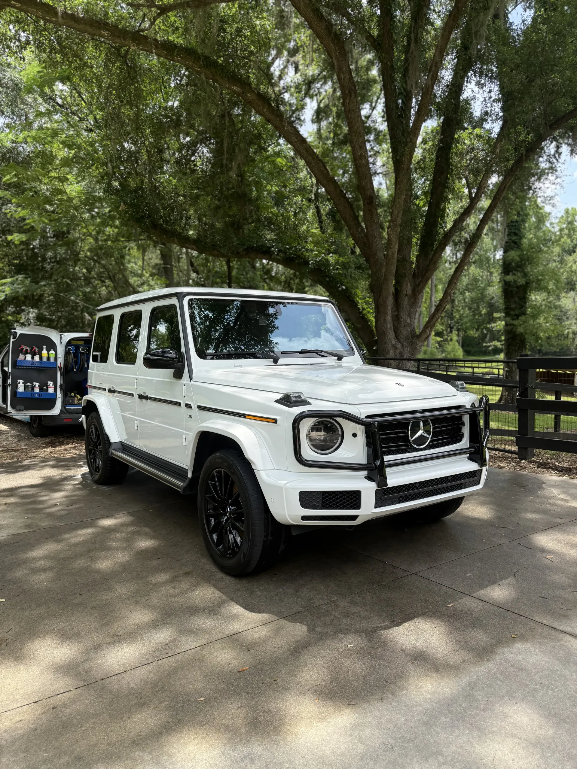 A white Mercedes-Benz G-Wagon SUV with black wheels parked on a paved driveway surrounded by trees.