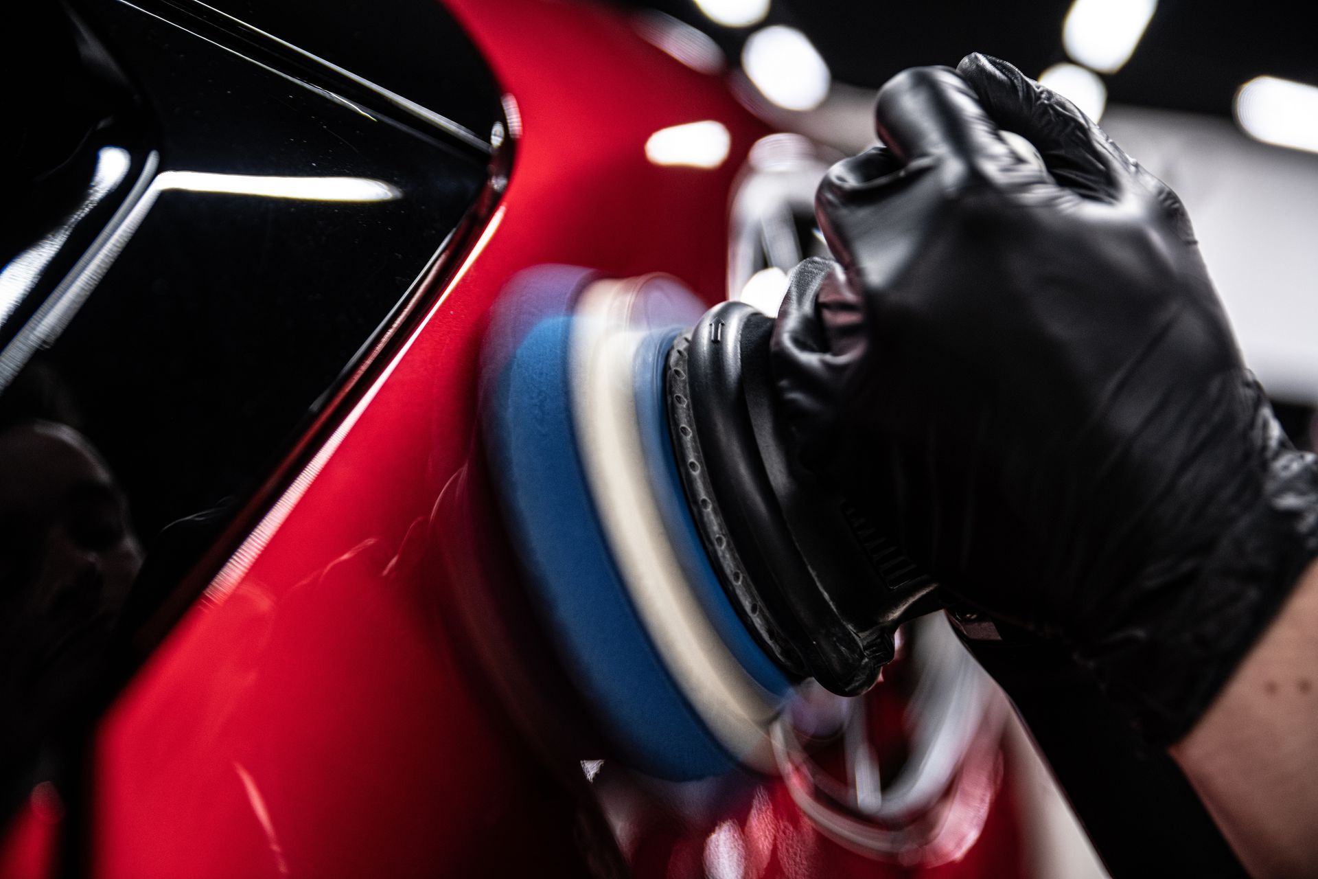 Close-up of a dark, metallic-painted car door panel with a small dent and scratch reflected in the overhead shop lights.