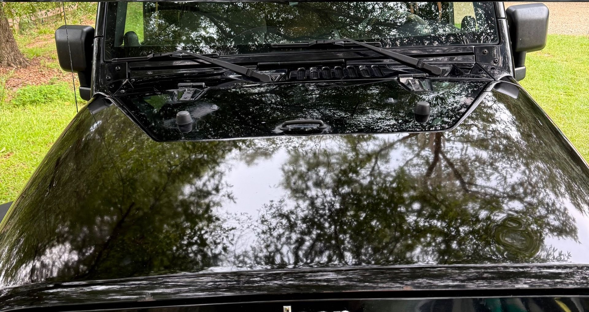 A high-angle view of a shiny black Jeep hood reflecting surrounding trees, with windshield wipers visible.