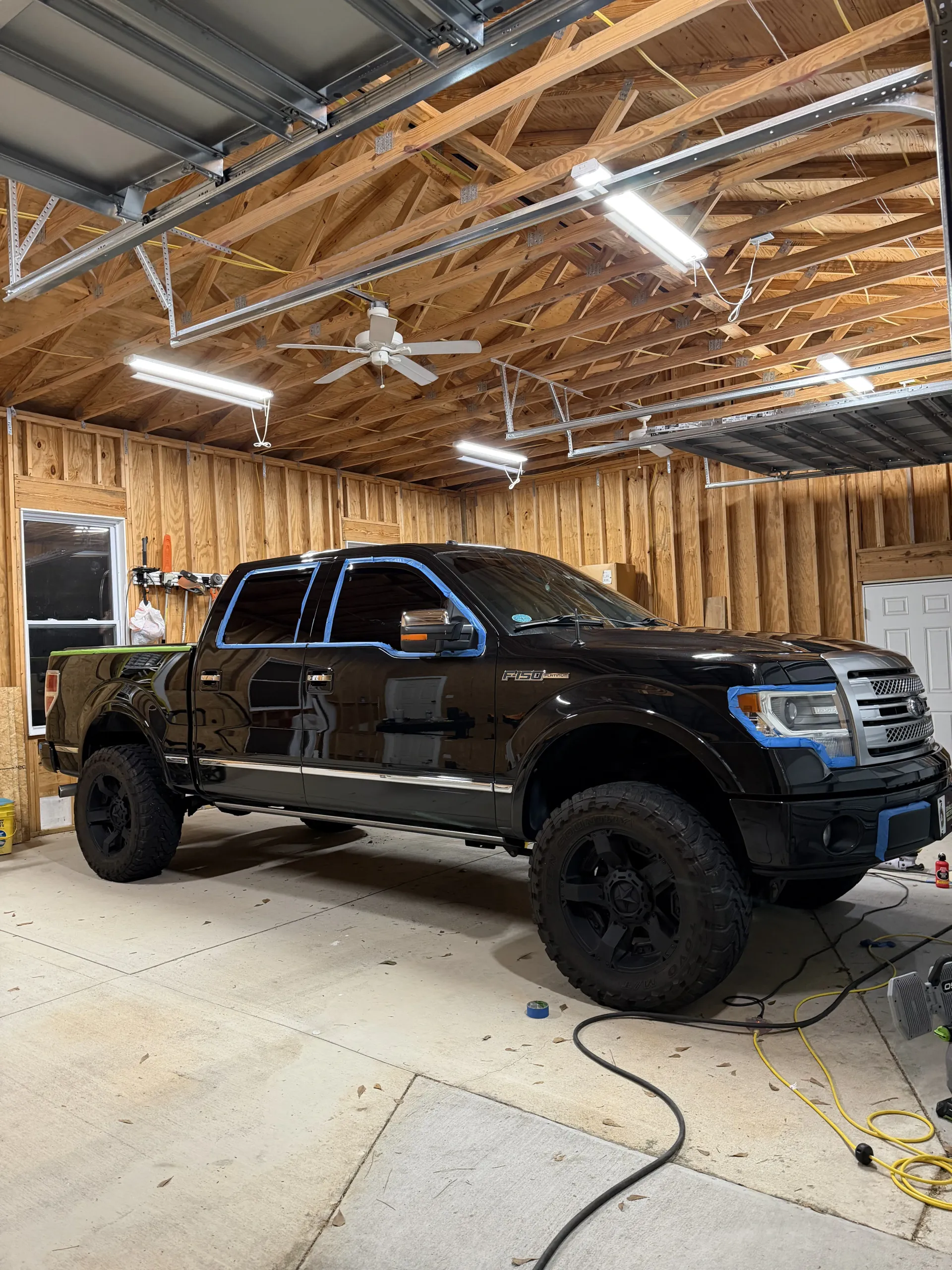 A lifted black pickup truck with blue painter's tape on its exterior is parked inside a wood-paneled garage.