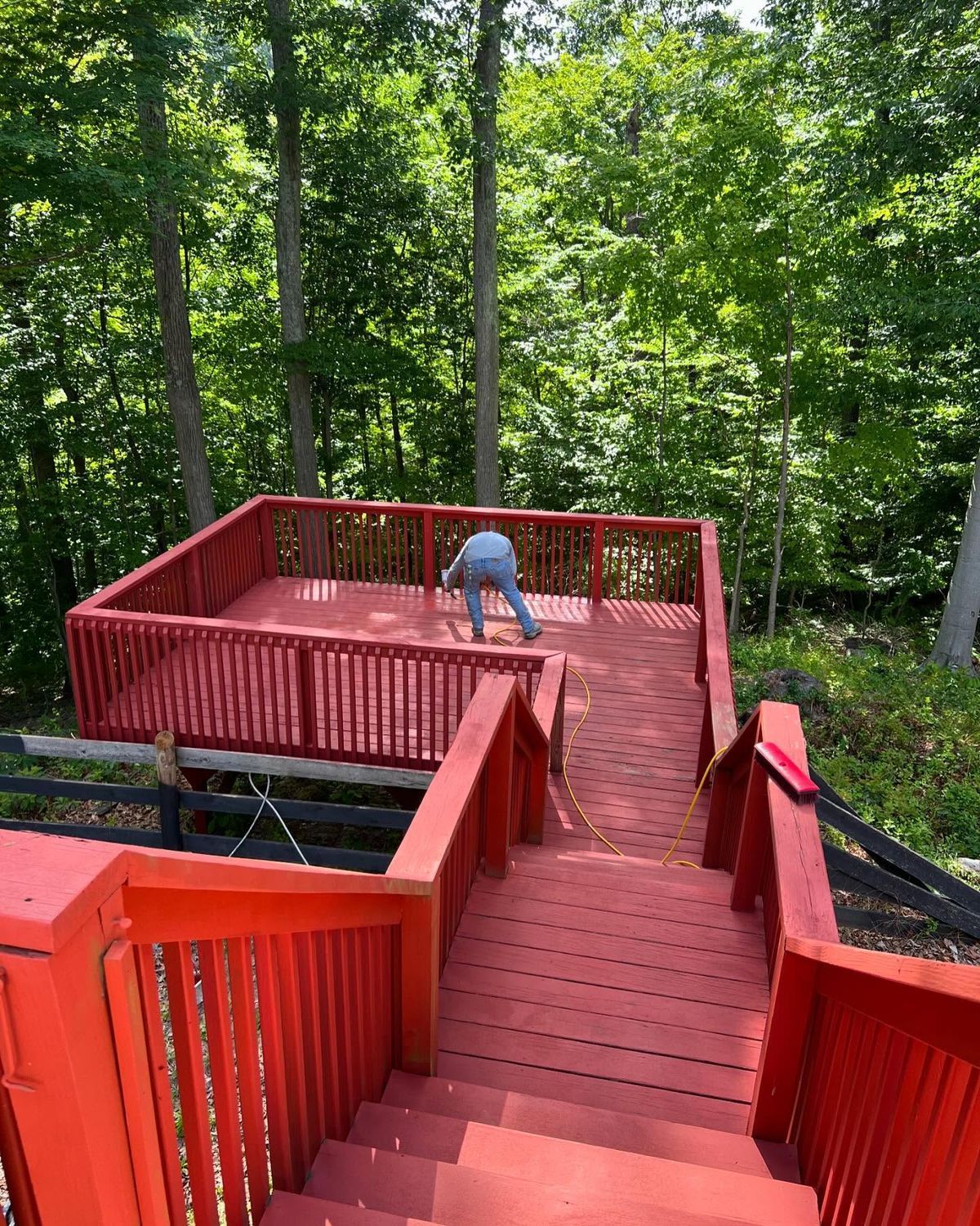Person painting a bright red wooden deck with stairs in a wooded setting.