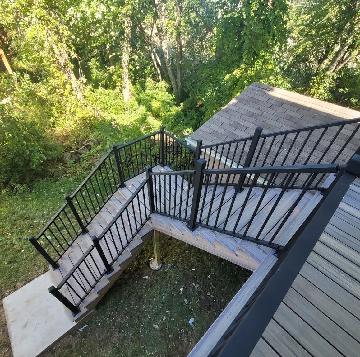 Staircase with black railing, leading from a deck to a grassy yard, surrounded by trees.