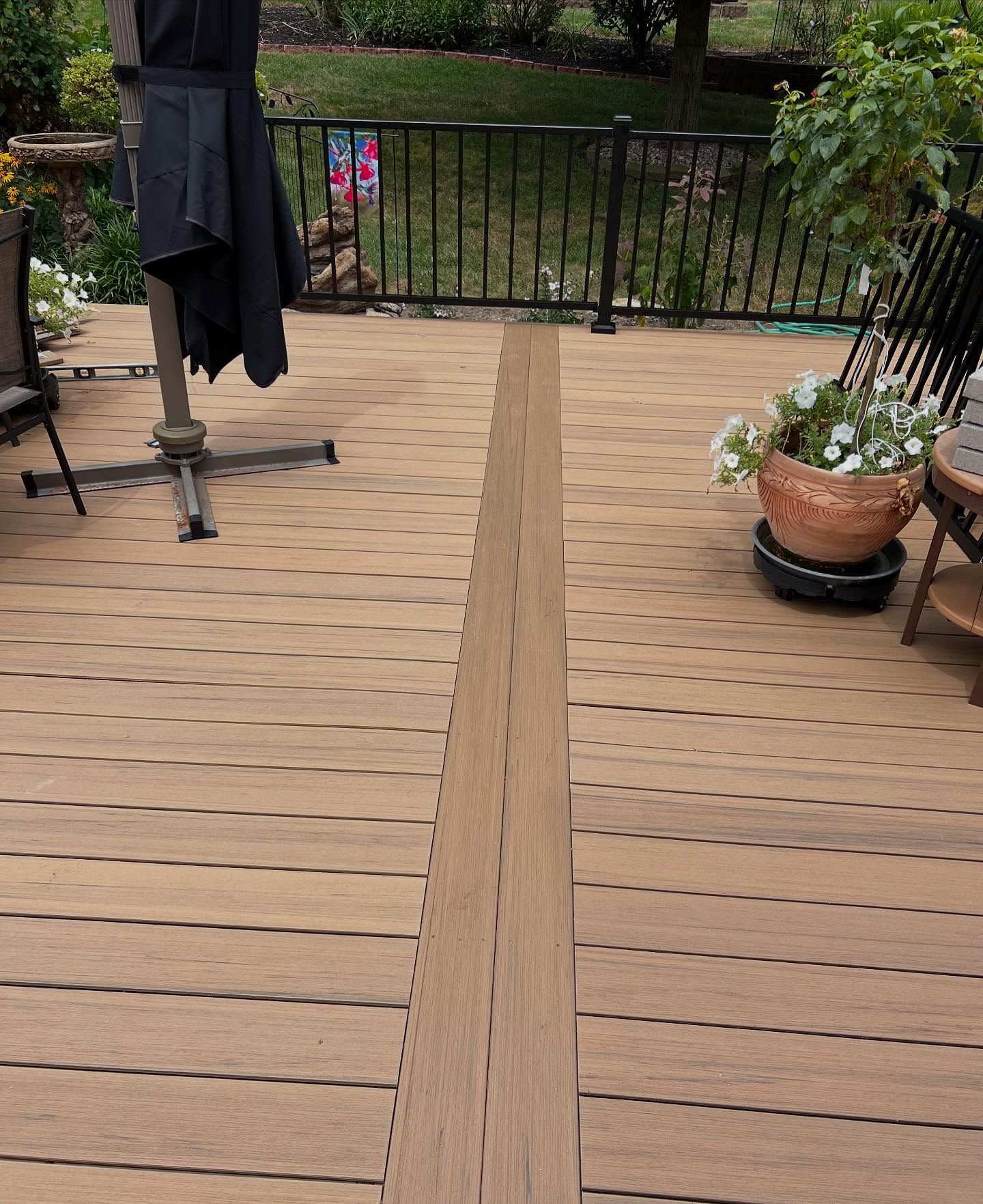 Wooden deck with a central brown strip; umbrella, plants, and railing in the background.