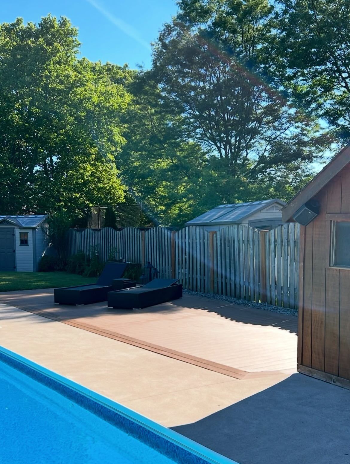 Poolside scene: blue pool edge, tan deck, two black lounge chairs, wooden shed, green trees, blue sky.