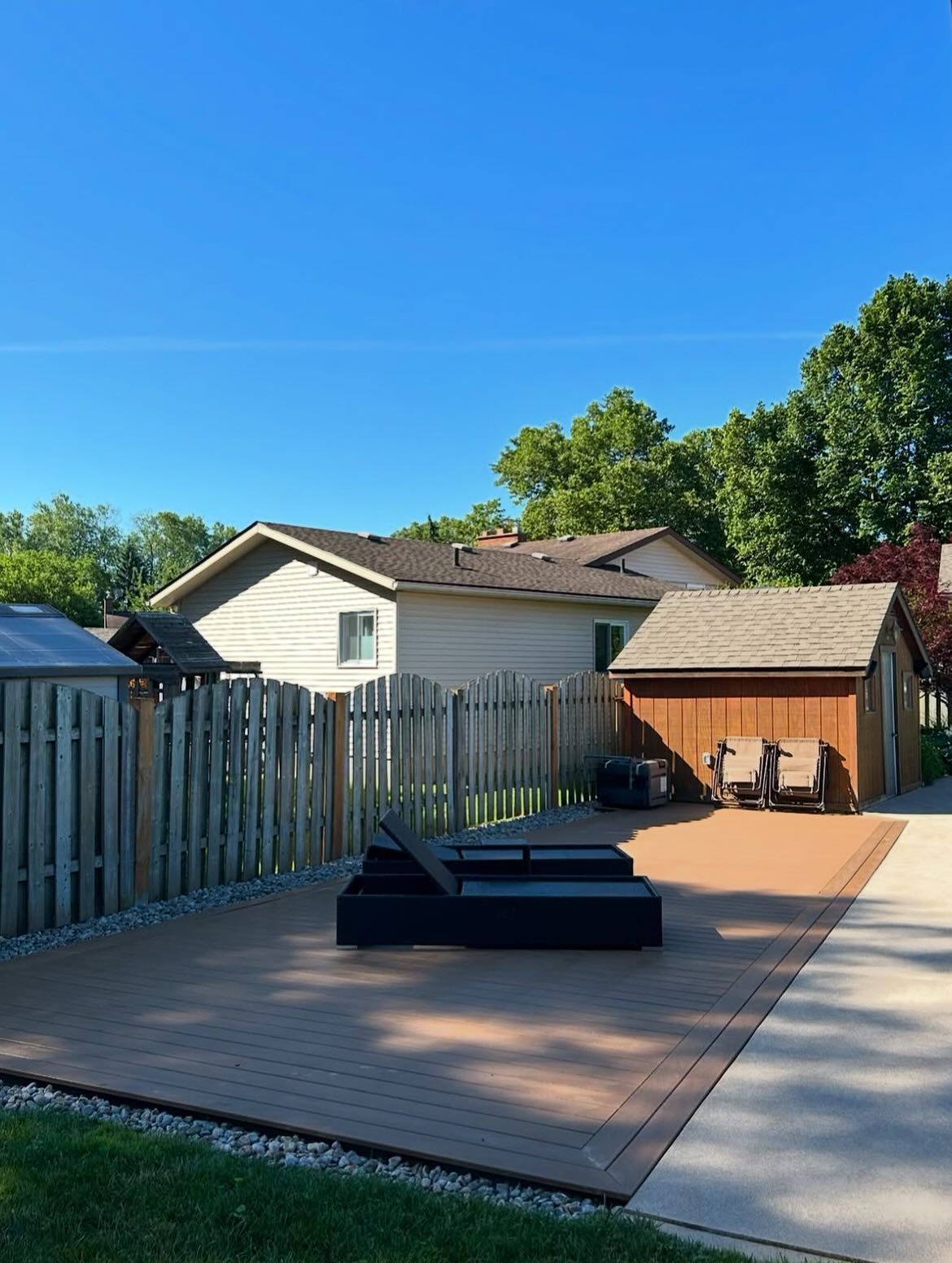 Backyard with a composite deck, lounge chairs, wooden shed, fence, and house under a blue sky.
