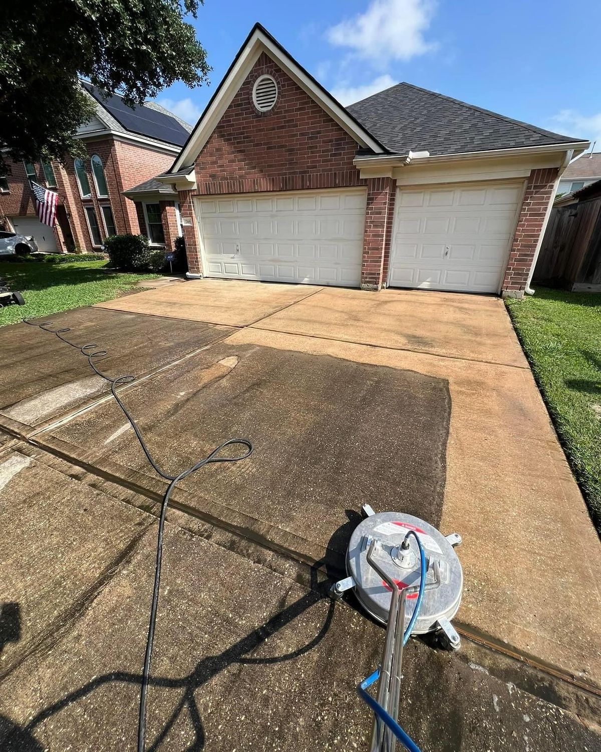 A person pressure washing a driveway in front of a brick house, cleaning a section of the concrete.