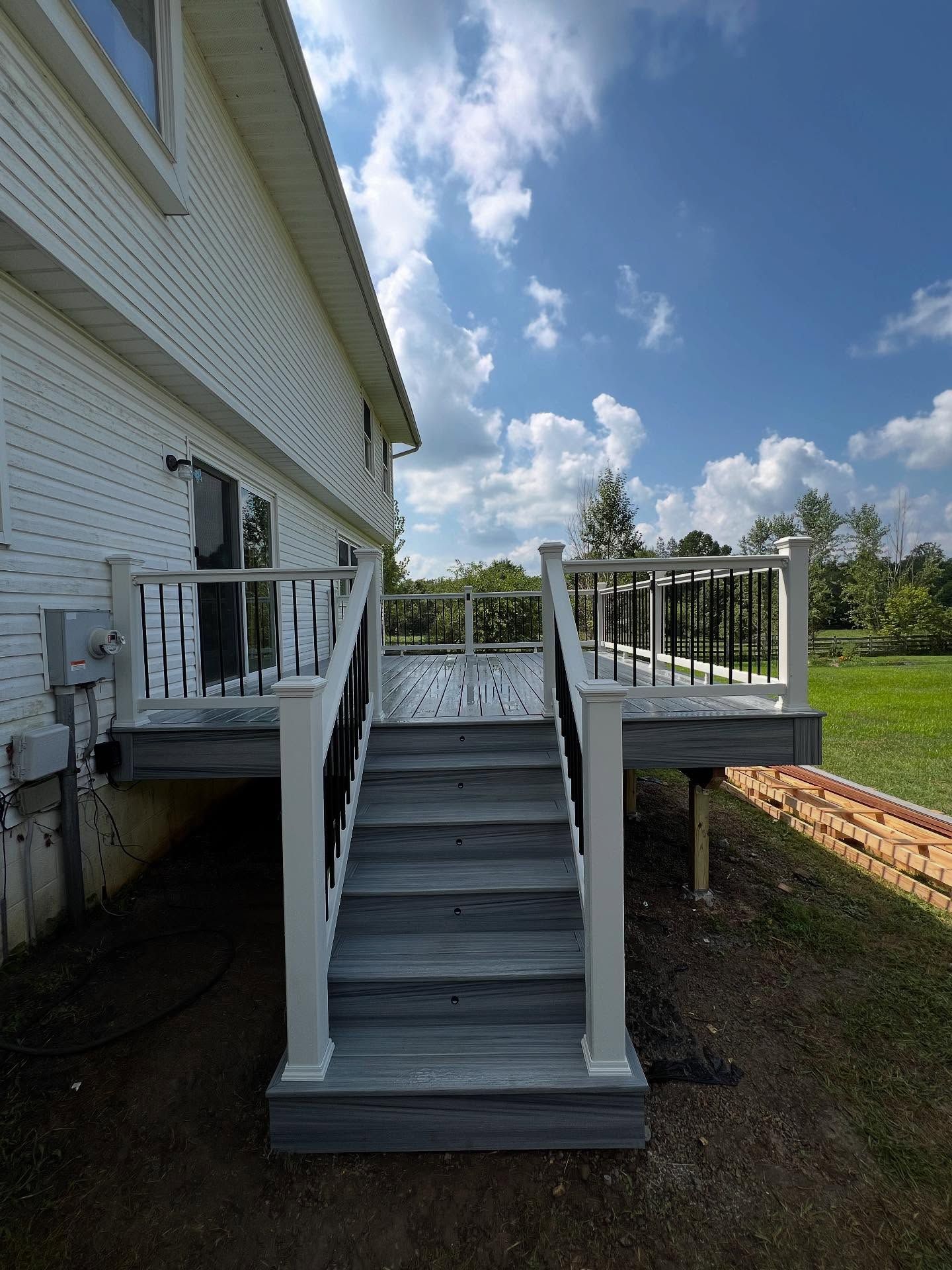 Grey deck with white and black railing and steps leading up to a white house on a sunny day.