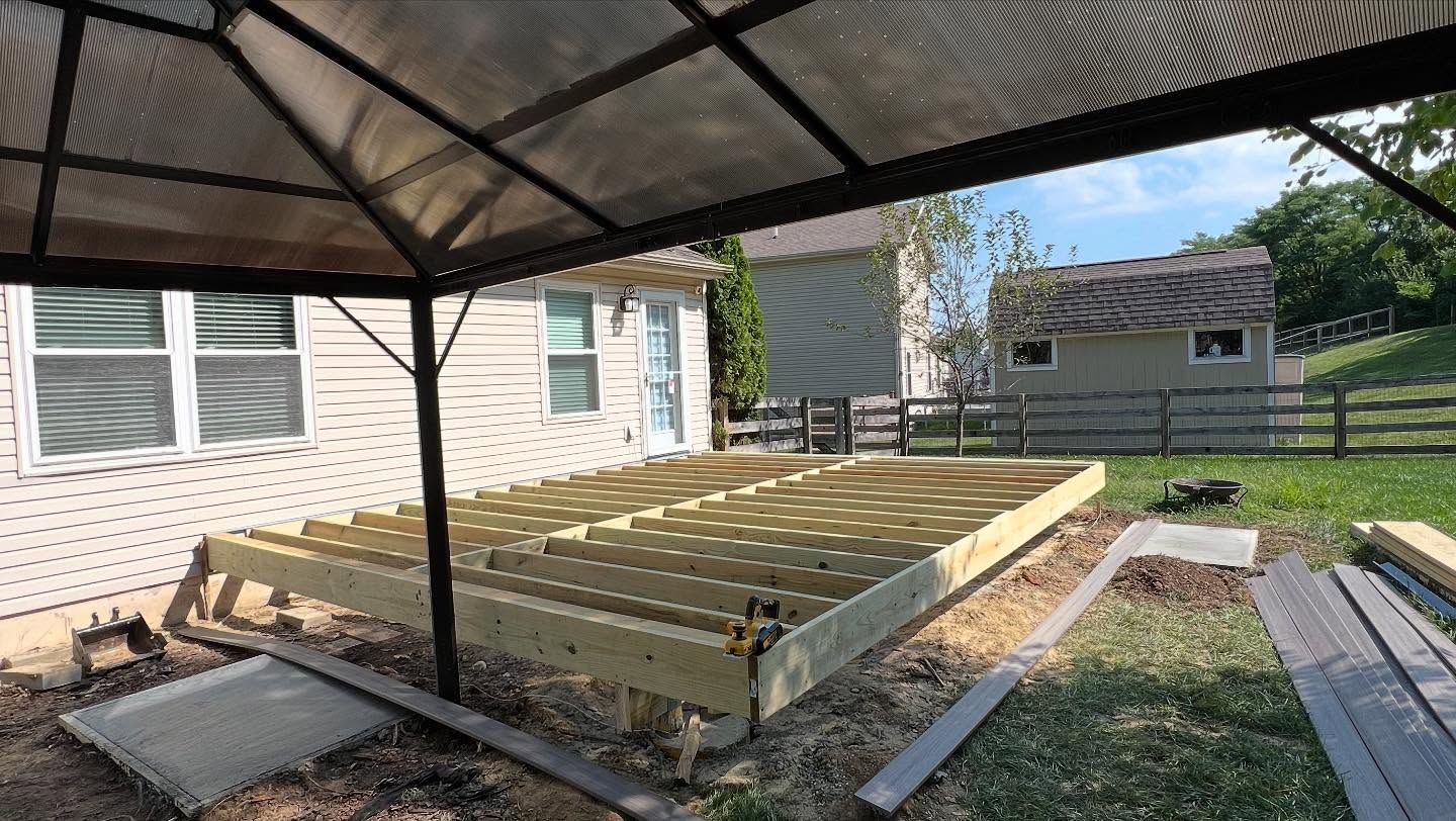 A deck in progress, wooden beams, dirt, gazebo frame, house, small shed, and green grass.