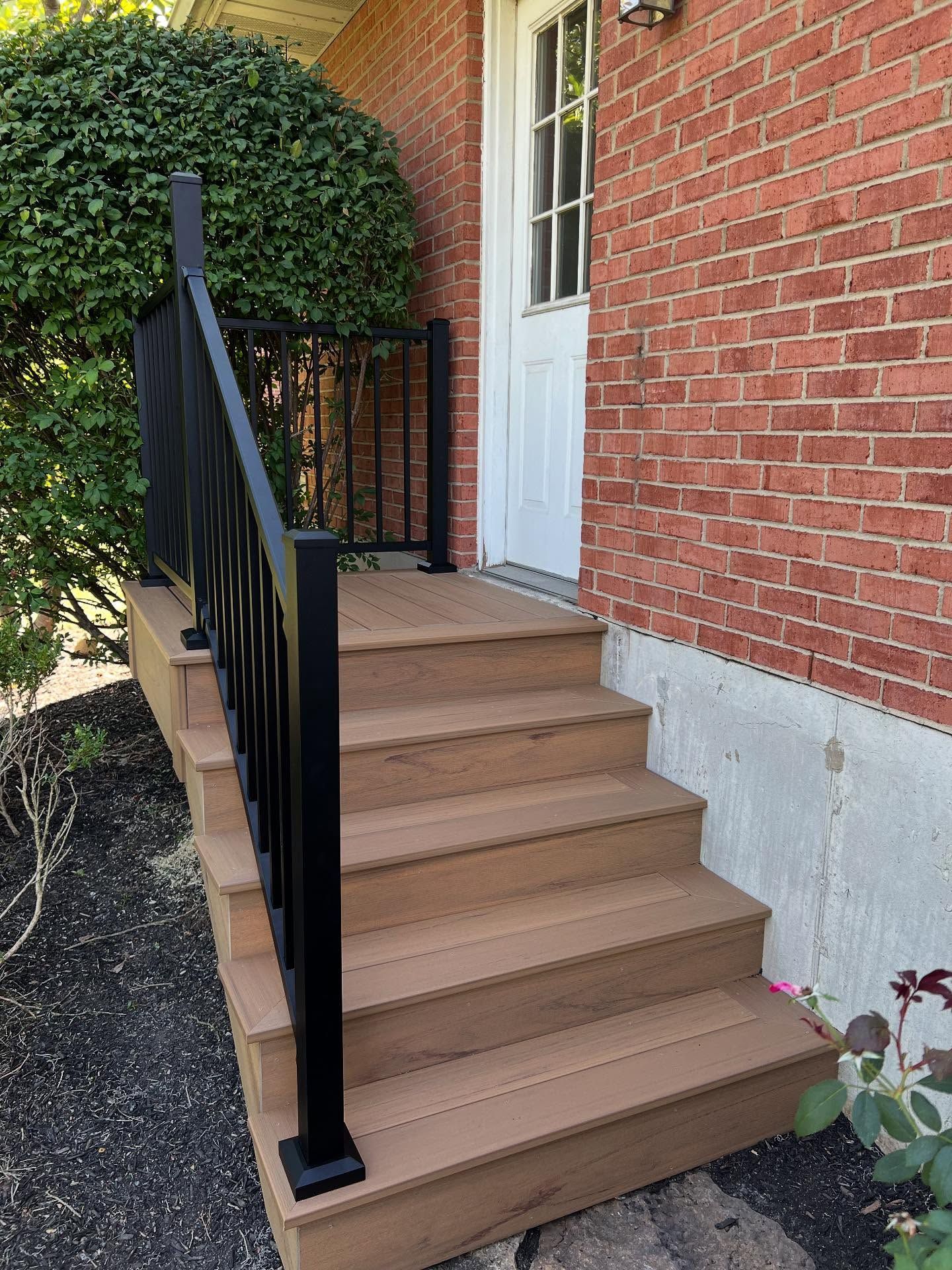 Steps leading to a doorway with black railing, against a brick wall and a bush.