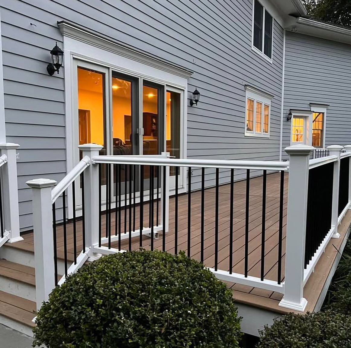 A deck with white railings and black spindles adjoins a gray house with a sliding glass door; lit interior visible.