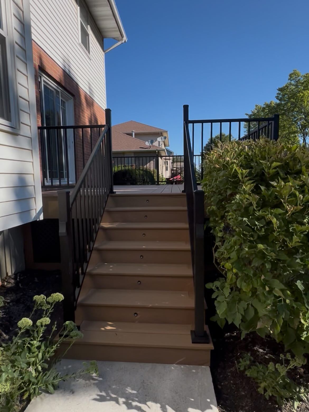 Wooden stairs leading to a deck, with black railing and a house on a sunny day.