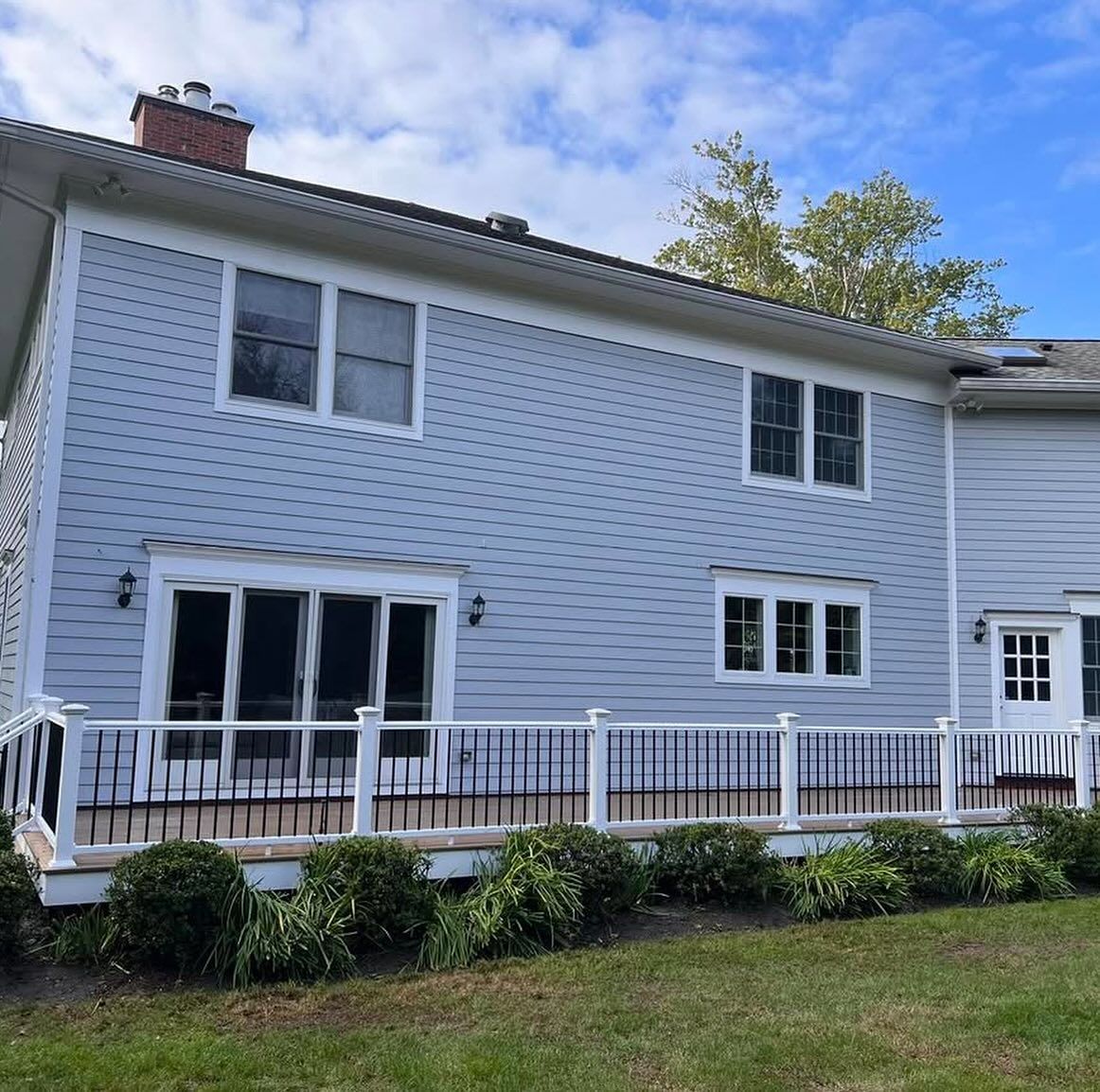 Two-story blue house with a deck, white railing, and windows. Green bushes and grass in front.