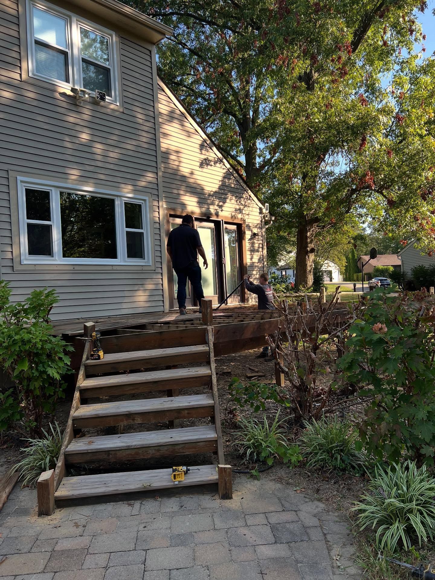 A person walks toward a house with a deck and stairs, alongside a garden with trees on a sunny day.