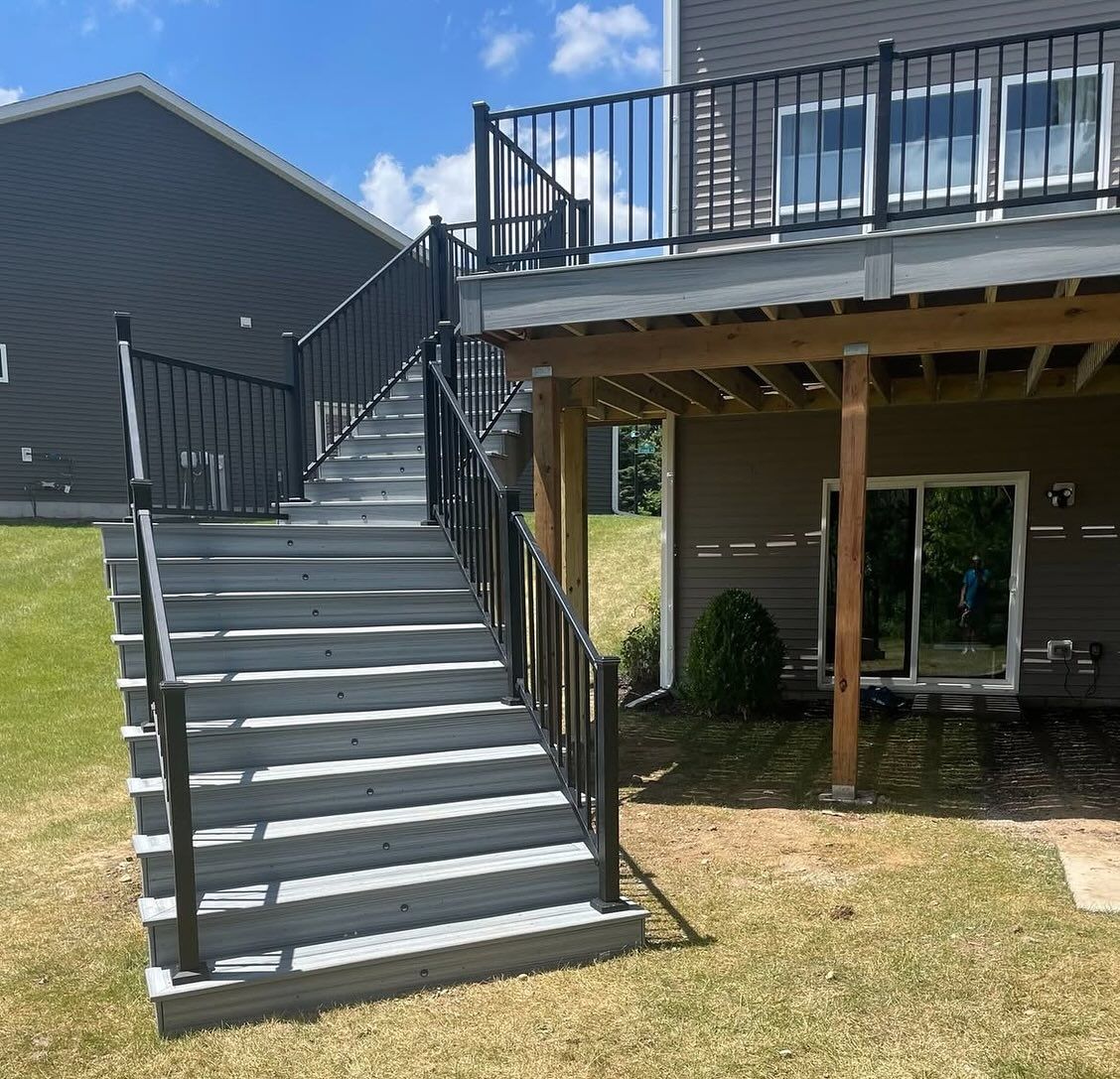 Outdoor deck with steps, black railing, grey wooden stairs, and a yard with grass and a house.