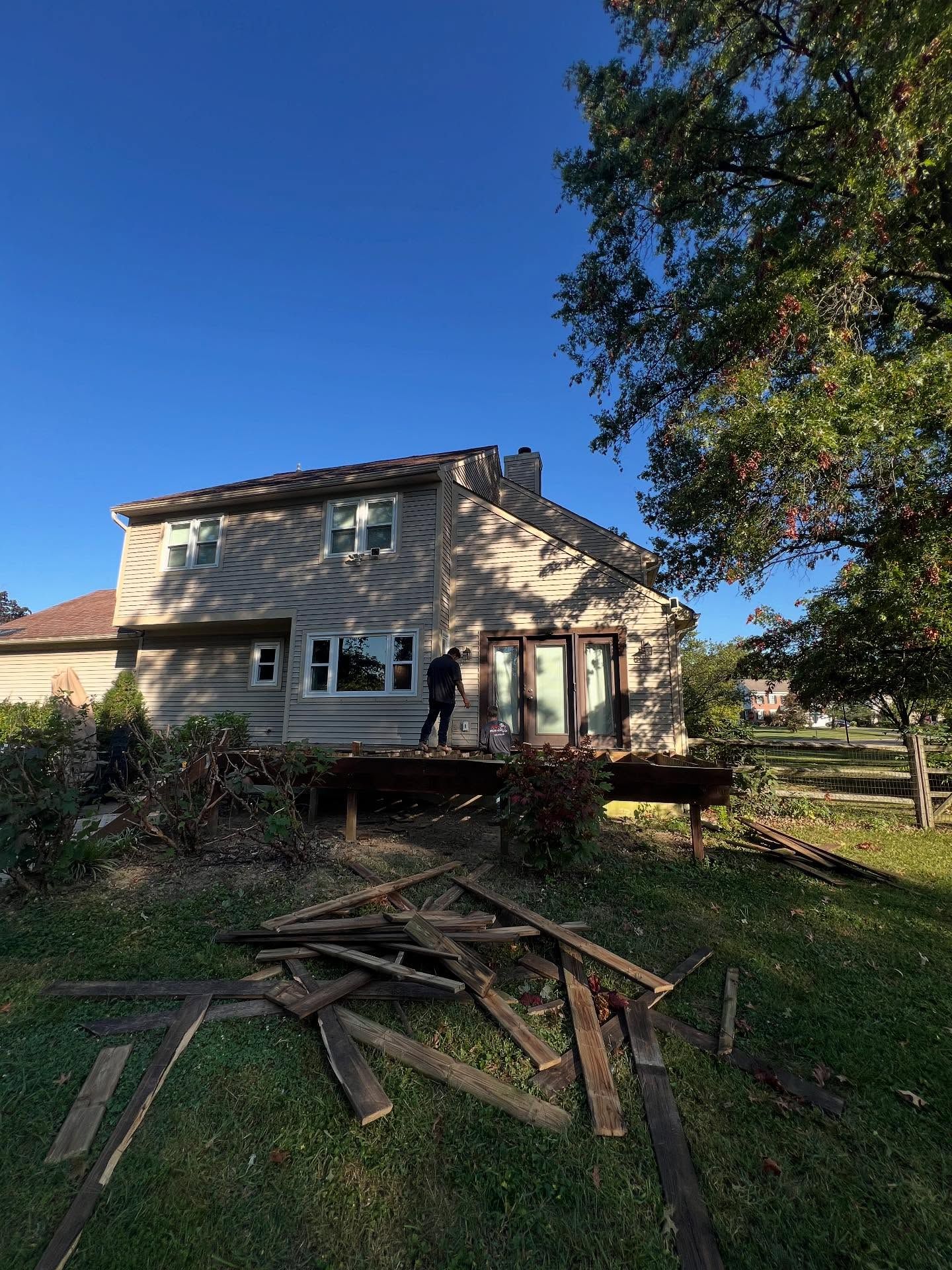 House with a person on the deck, debris on the ground, and a clear blue sky.