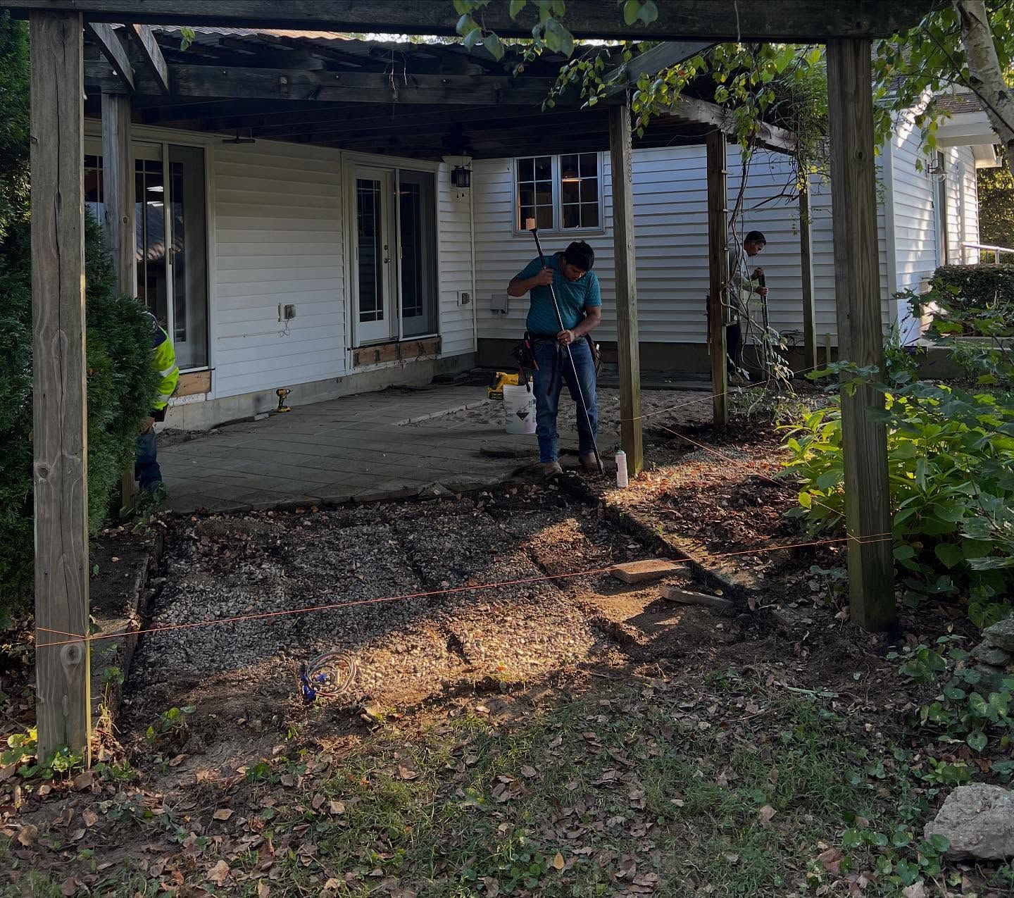 Two people working in a backyard under a wooden pergola. One digs with a shovel, the other is farther back.