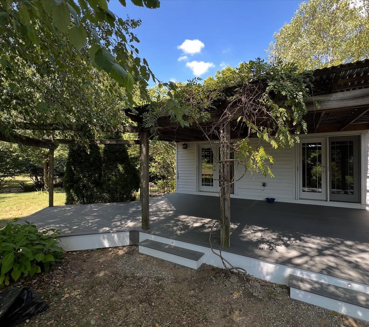 A white house with a porch, pergola covered in vines, blue sky.