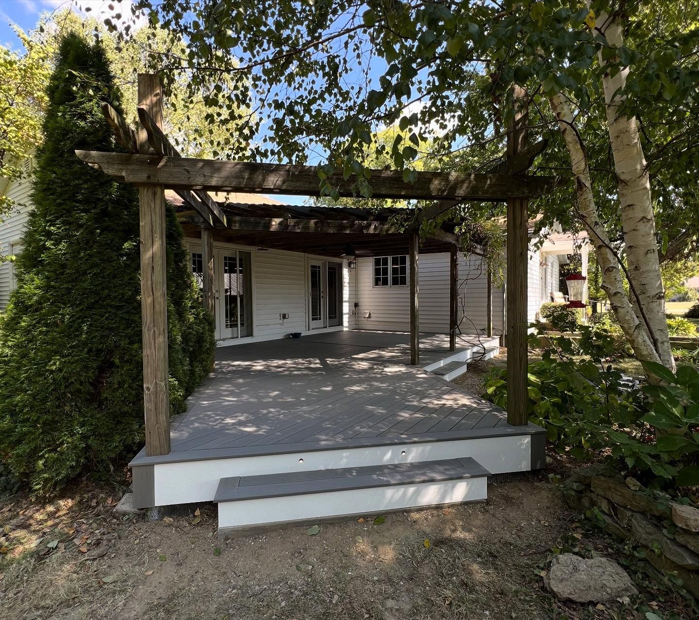 Wooden pergola over a gray deck with steps, attached to a white house, outdoors.