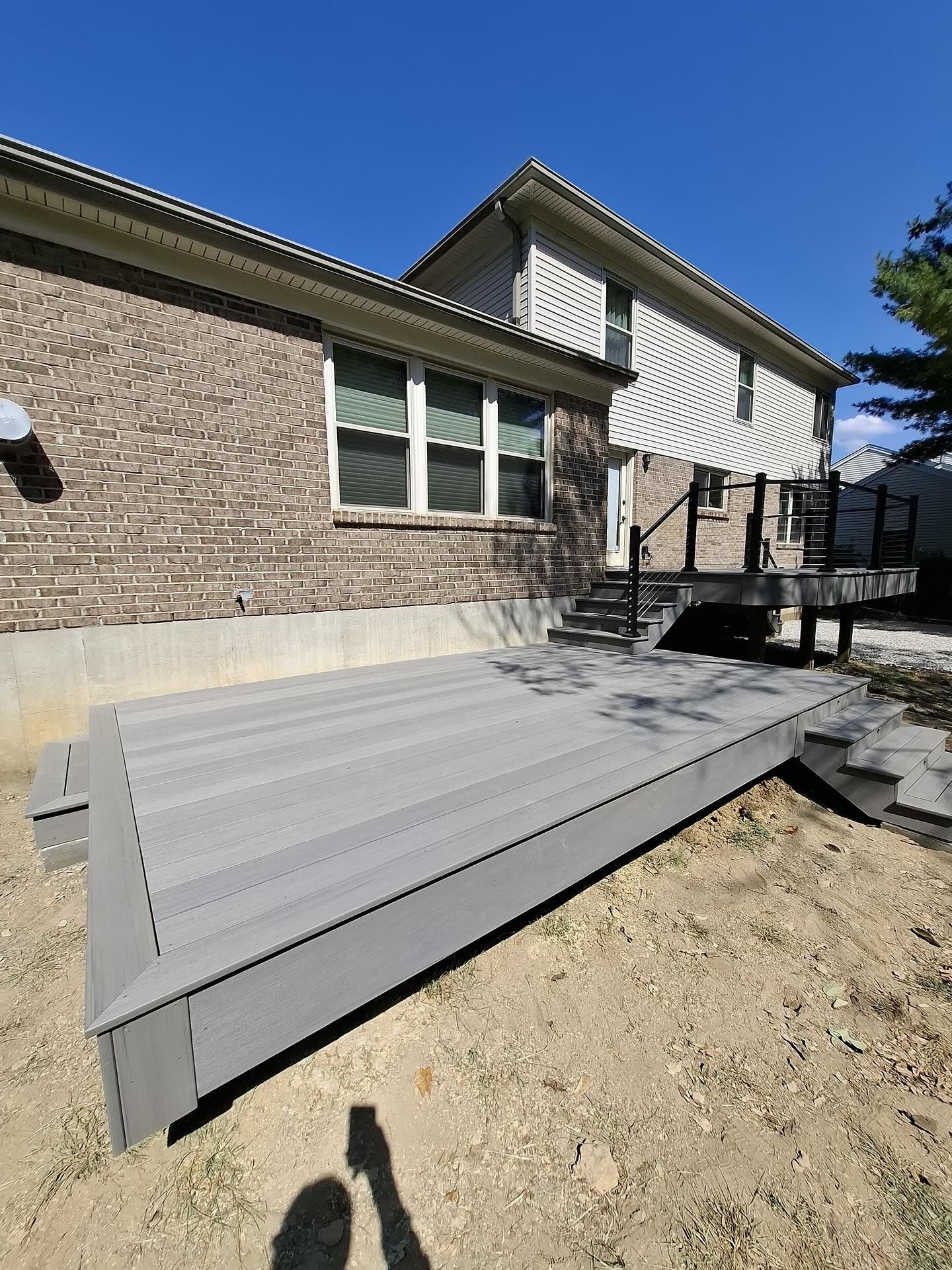 Gray composite deck with stairs, attached to a two-story house with brick and siding, sunny outdoors.