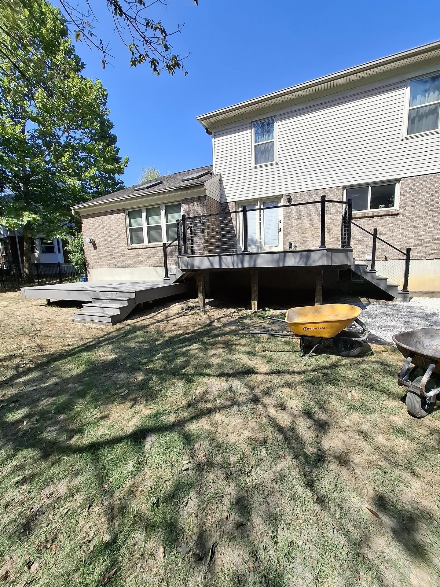 Backyard deck with stairs, overlooking grassy yard, yellow wheelbarrow, and two-story brick house.