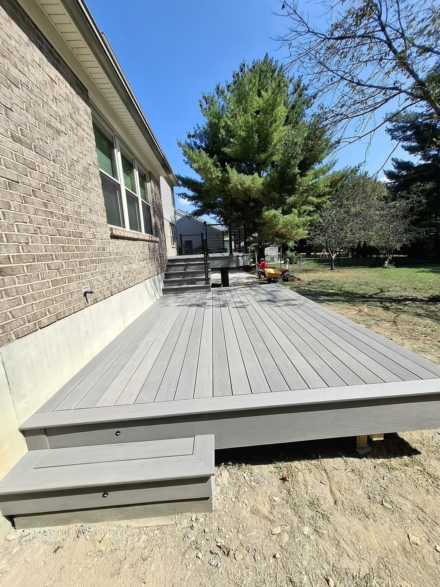 Gray composite deck with stairs next to a brick house and a tall tree on a sunny day.
