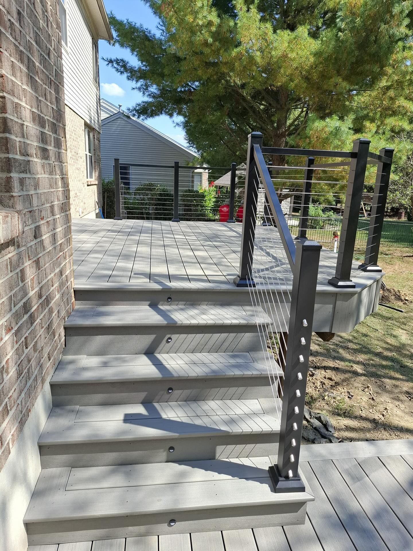 Composite deck with stairs, metal railing, and brick wall, overlooking a yard with a tree.