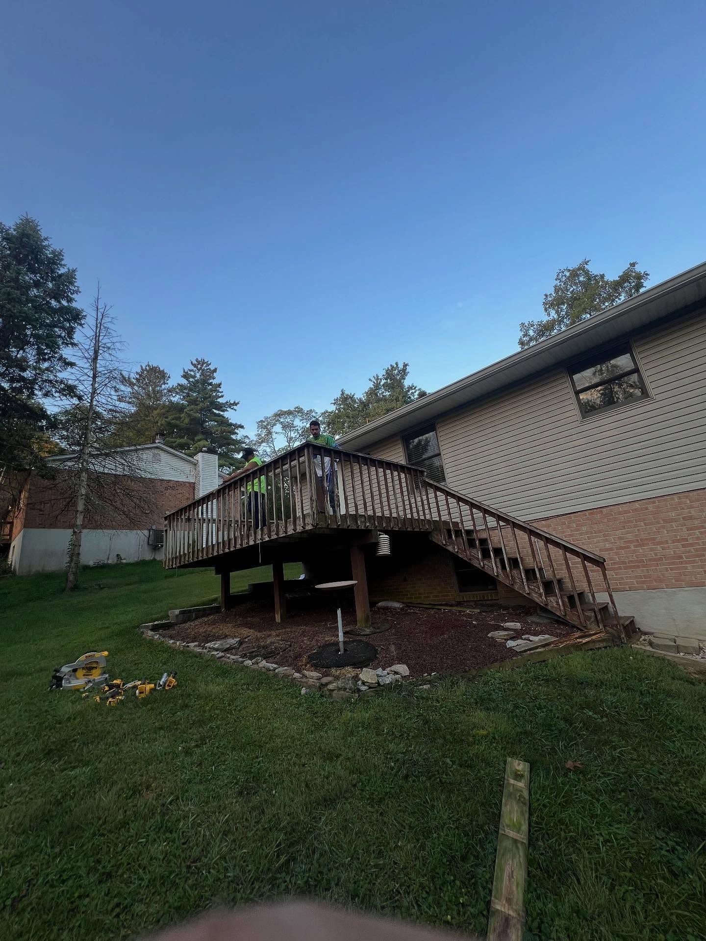 A wooden deck on a grassy hill leads to a brick house. A person stands on the deck.