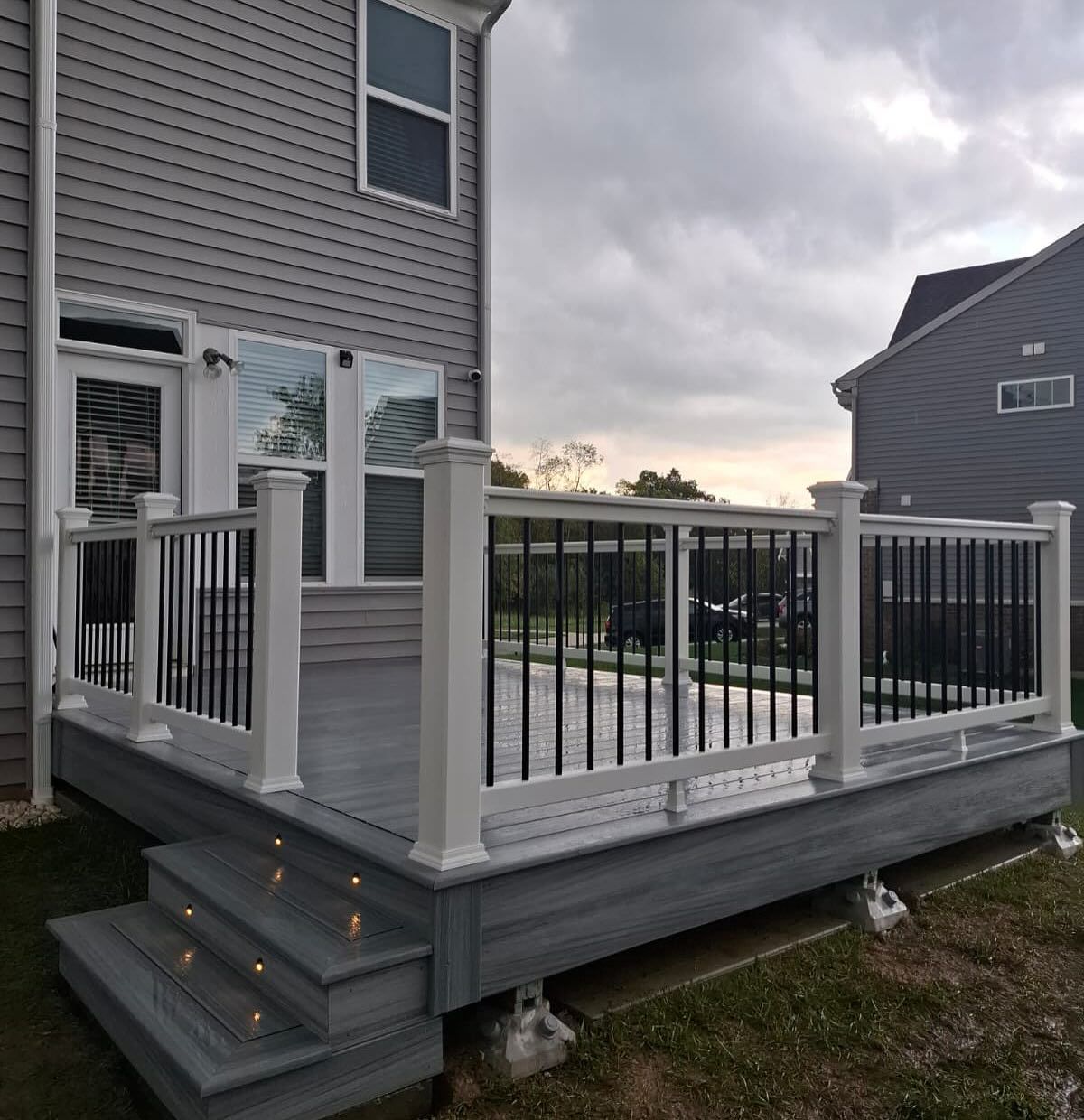 Gray deck with white posts, black railings, and steps, attached to a gray house, under a cloudy sky.