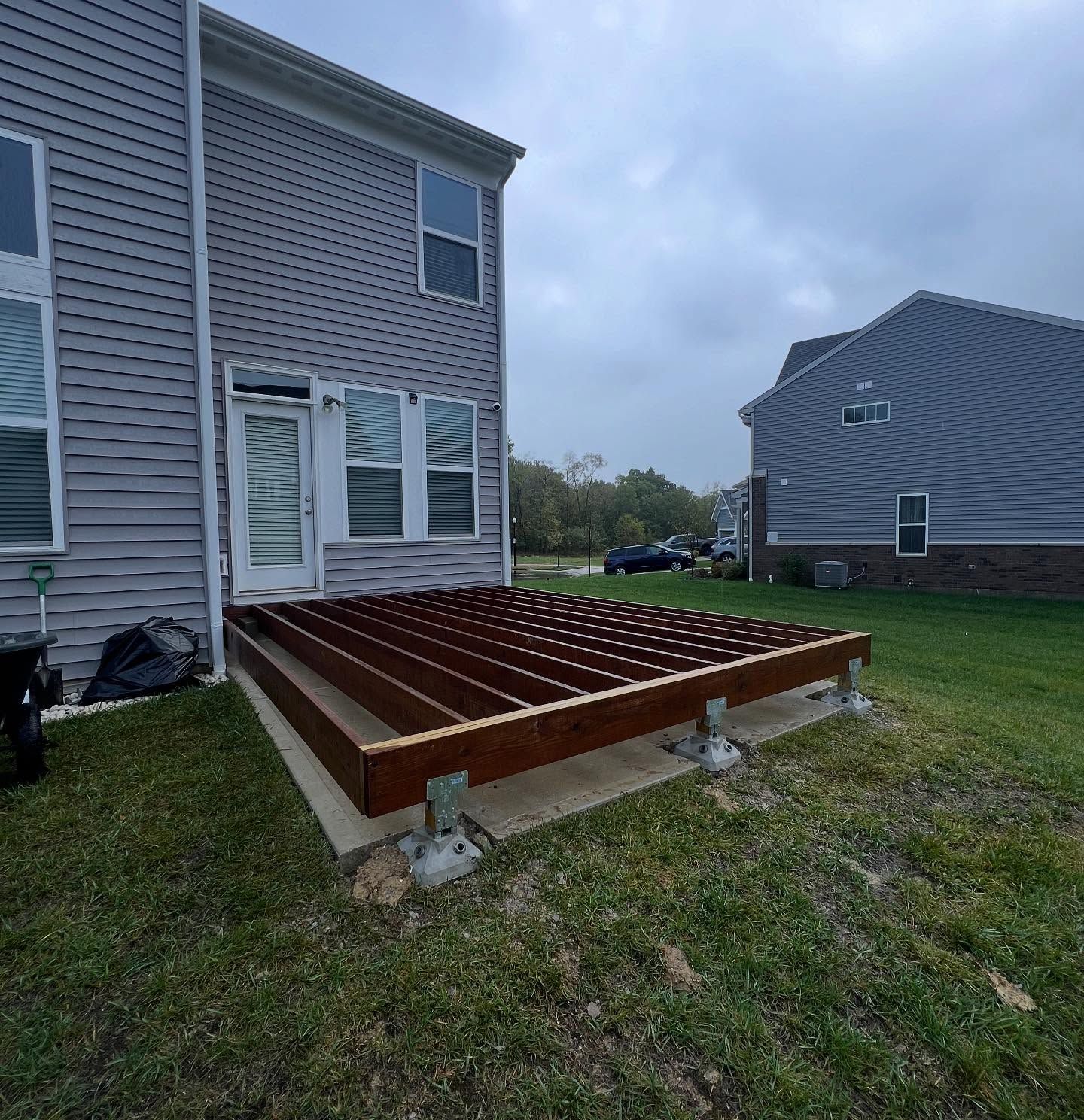A wooden deck under construction outside a gray house. Joists are laid on concrete blocks. Green grass surrounds.