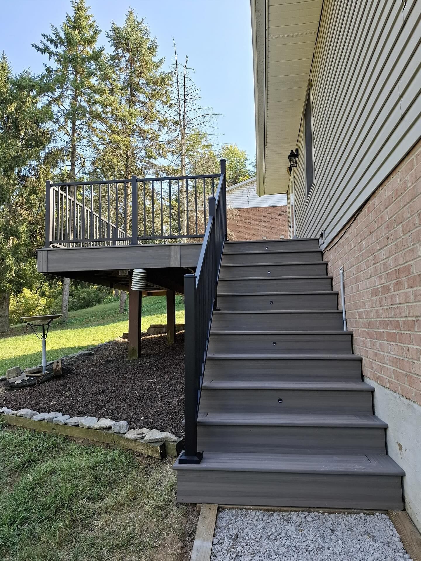 Grey deck with stairs leading down to a yard with dark railing and light siding building.
