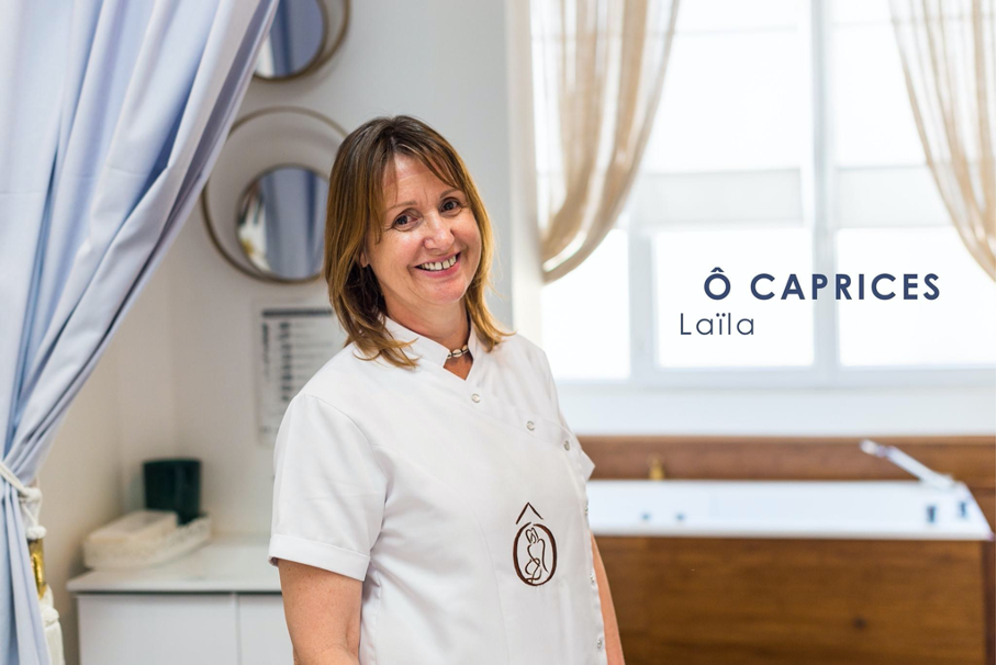 Femme, Laila, en uniforme blanc souriant à la caméra dans un décor de type spa.